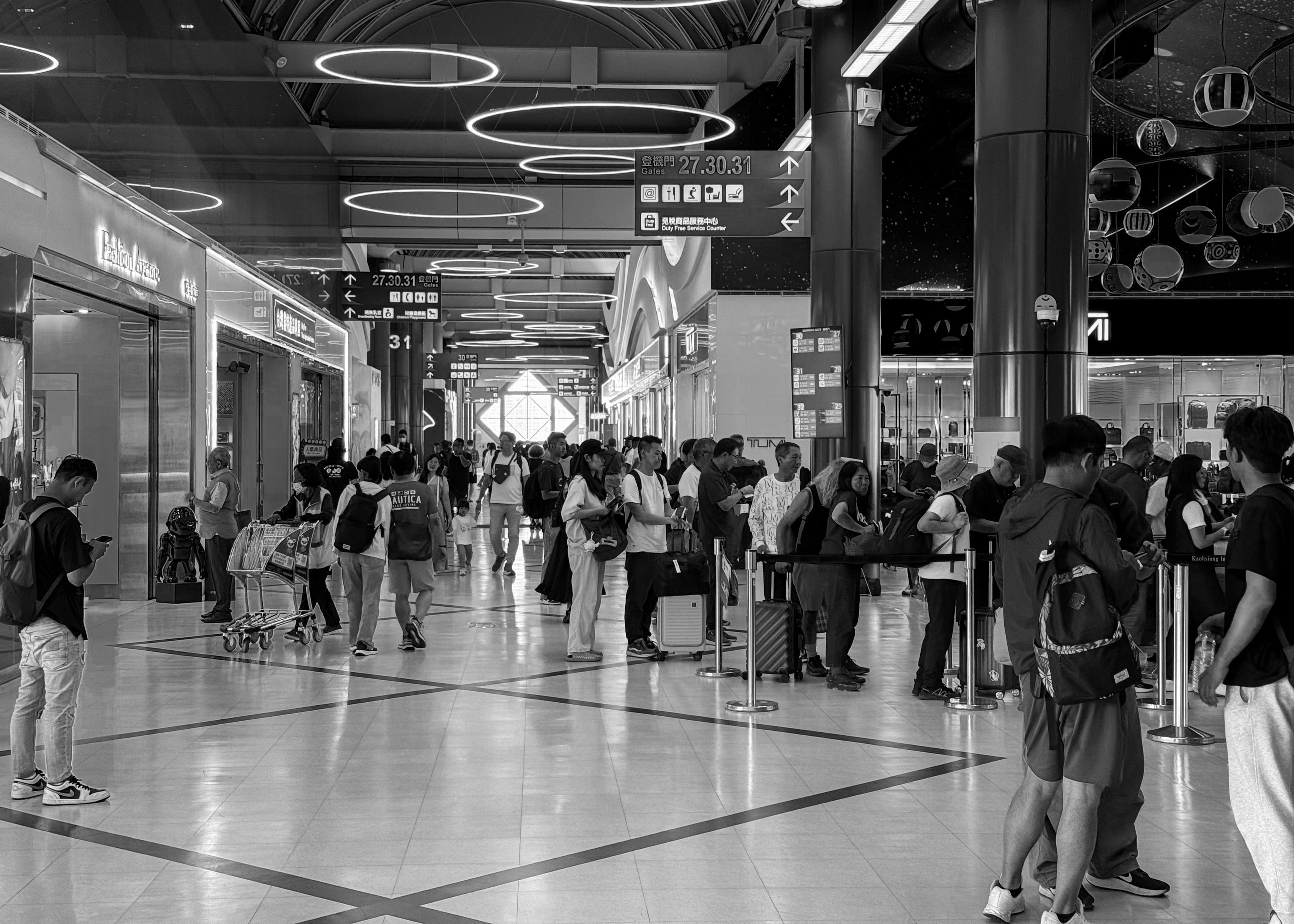 Crowd of travelers standing in line at an airport check-in or security area, with some waiting and others using their phones.