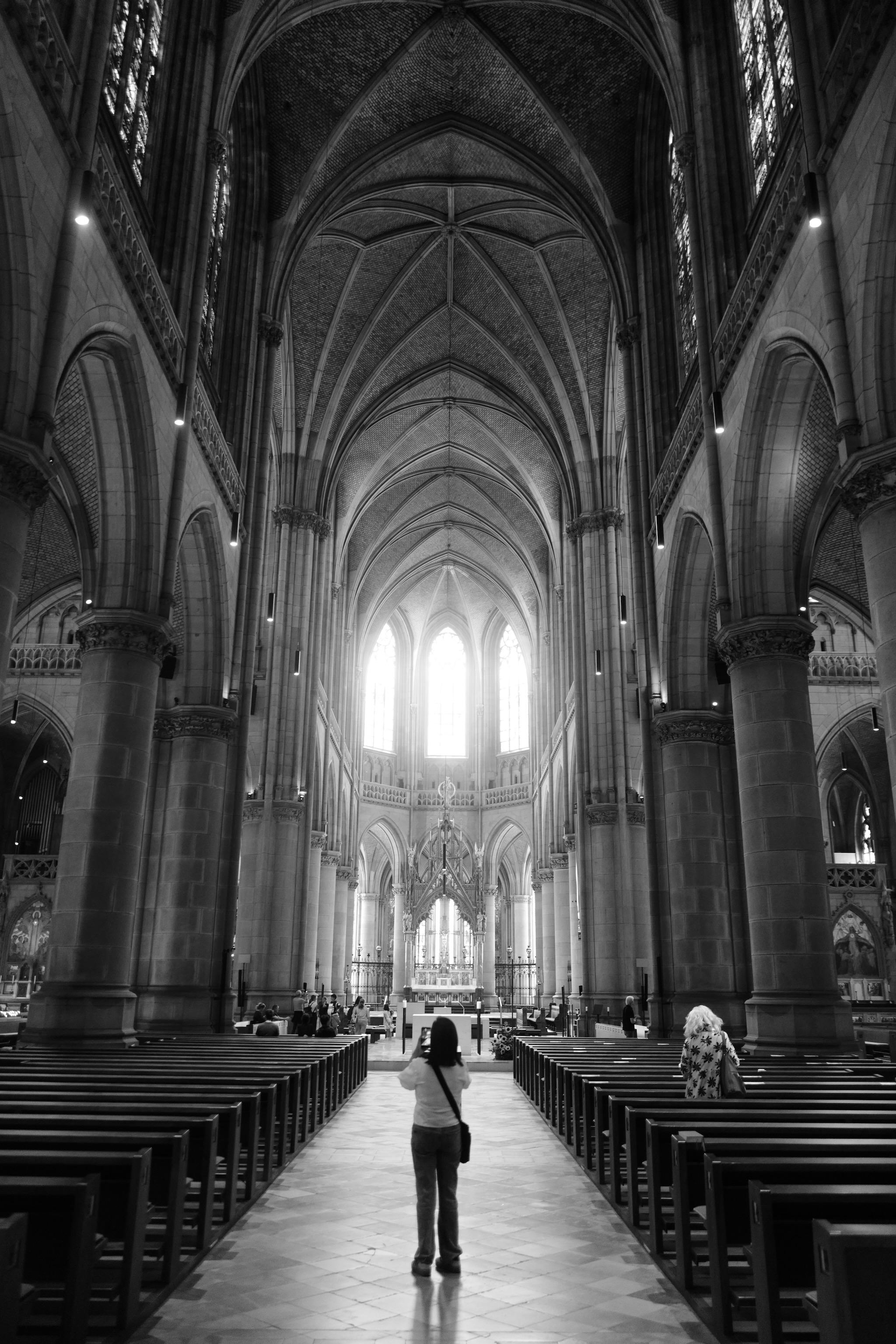 Picture of the interior of a gothic church. The building is the New Cathedral of Linz, a neo-gothic style church finished in 1902. 
In front of the photo is a woman, taking a picture of the building. In the background is the space of the altar and three gothic windows with bright light coming in. On the upper side the rip vault is visible, also the pillars carrying the roof.
Picture is black and white.