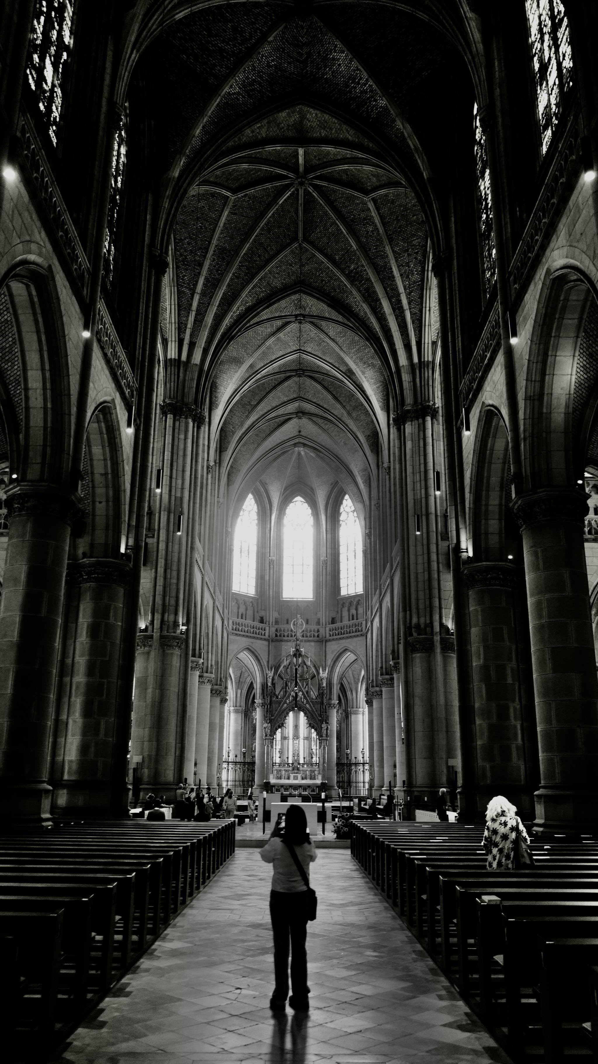 Picture of the interior of a gothic church. The building is the New Cathedral of Linz, a neo-gothic style church finished in 1902. 
In front of the photo is a woman, taking a picture of the building. In the background is the space of the altar and three gothic windows with bright light coming in. On the upper side the rip vault is visible, also the pillars carrying the roof.
Picture is black and white and edited to more darkness and contrast.