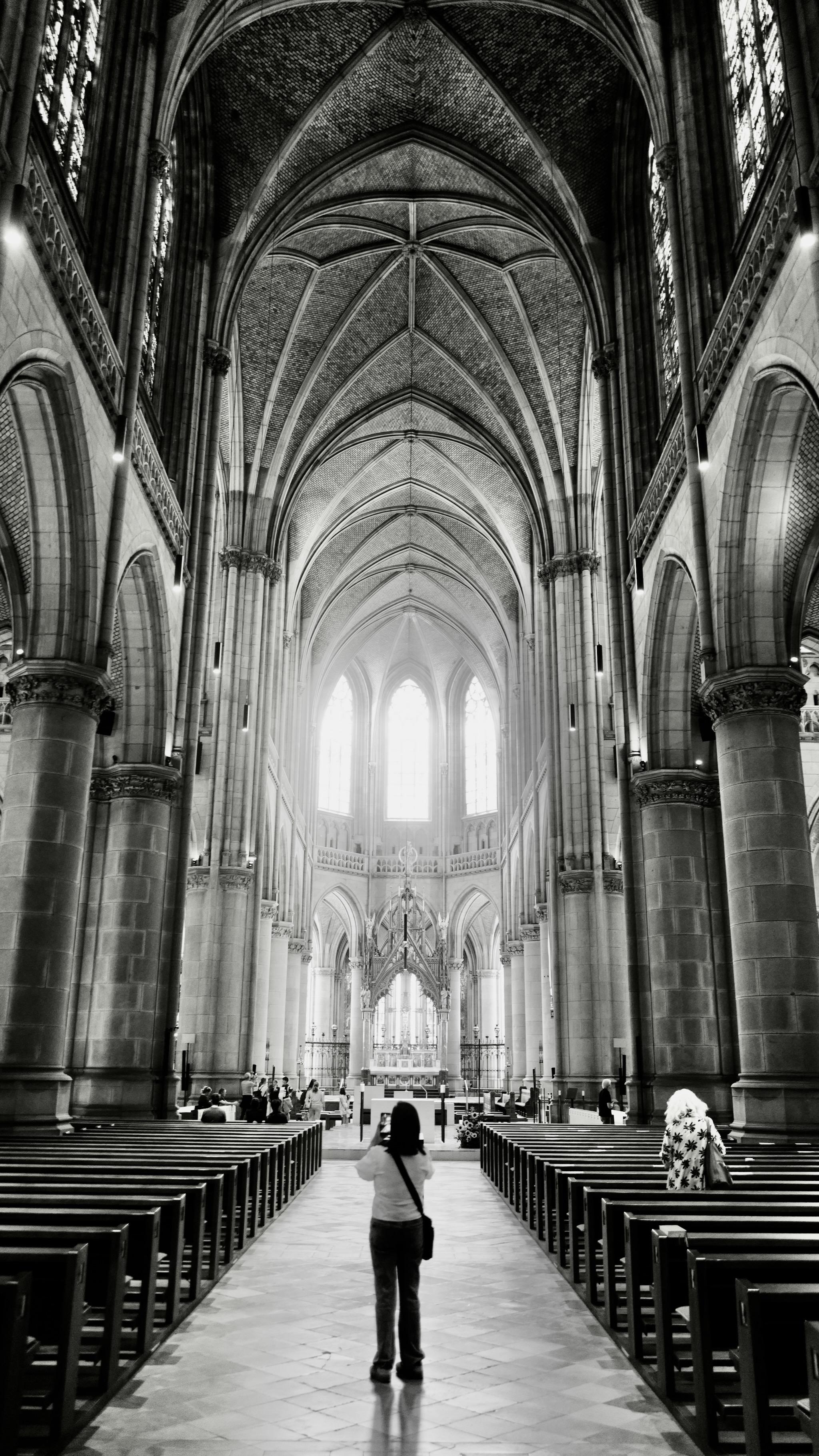 Picture of the interior of a gothic church. The building is the New Cathedral of Linz, a neo-gothic style church finished in 1902. 
In front of the photo is a woman, taking a picture of the building. In the background is the space of the altar and three gothic windows with bright light coming in. On the upper side the rip vault is visible, also the pillars carrying the roof.
Picture is black and white and edit to more brightness and highlights.