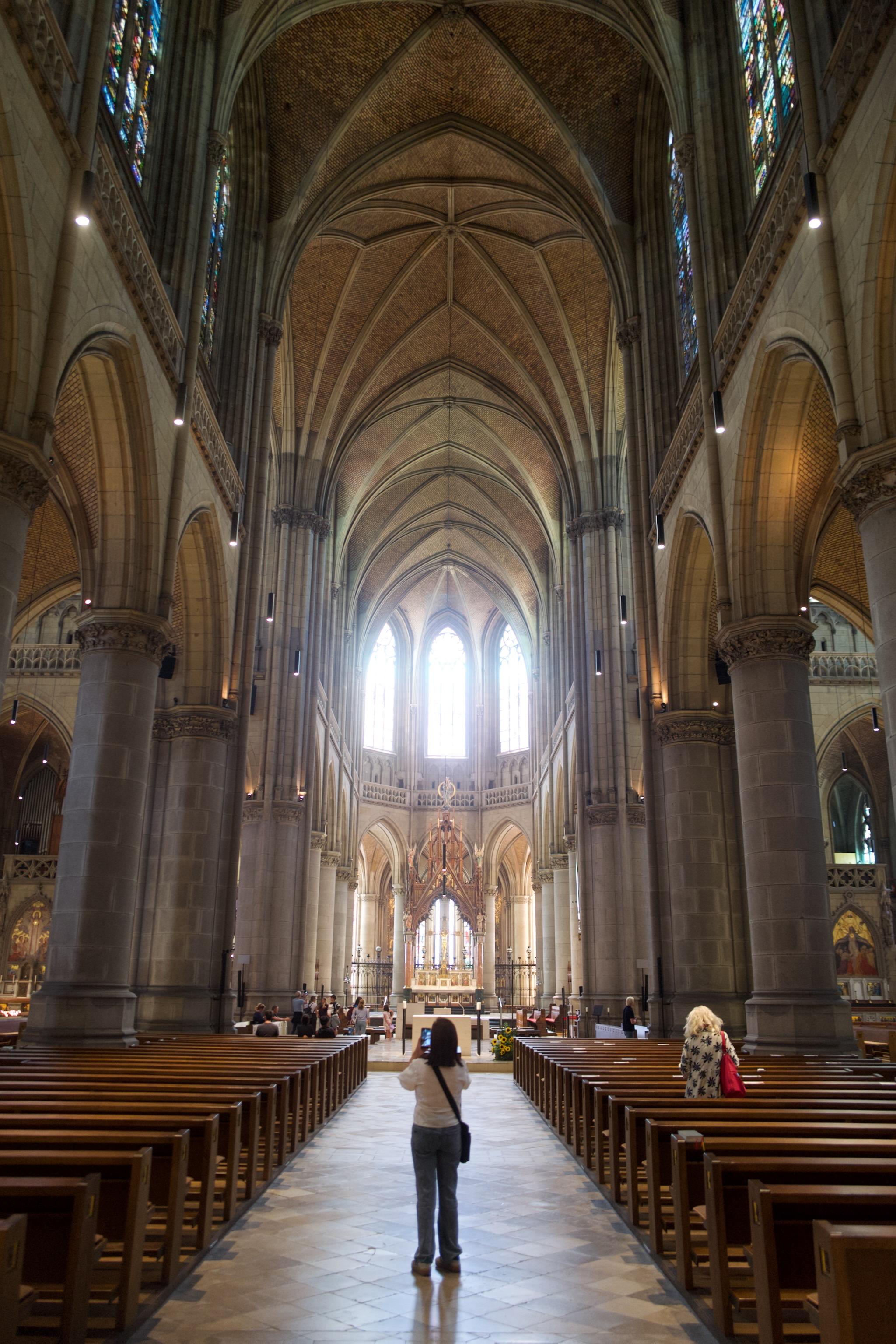 Picture of the interior of a gothic church. The building is the New Cathedral of Linz, a neo-gothic style church finished in 1902. 
In front of the photo is a woman, taking a picture of the building. In the background is the space of the altar and three gothic windows with bright light coming in. On the upper side the rip vault is visible, also the pillars carrying the roof.
Picture is colored.