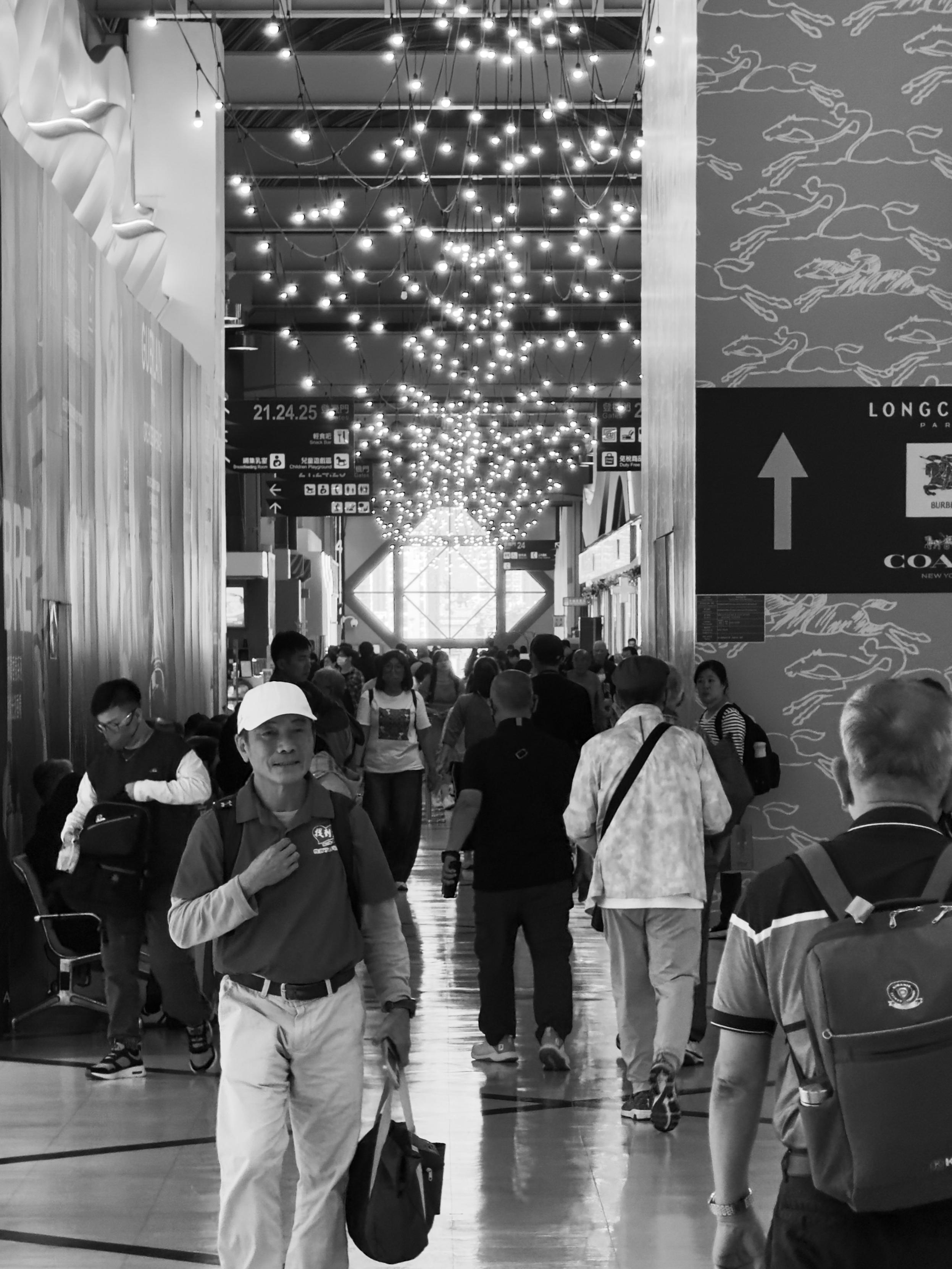 Crowded airport terminal with people walking, some carrying luggage, and decorative lights hanging from the ceiling.