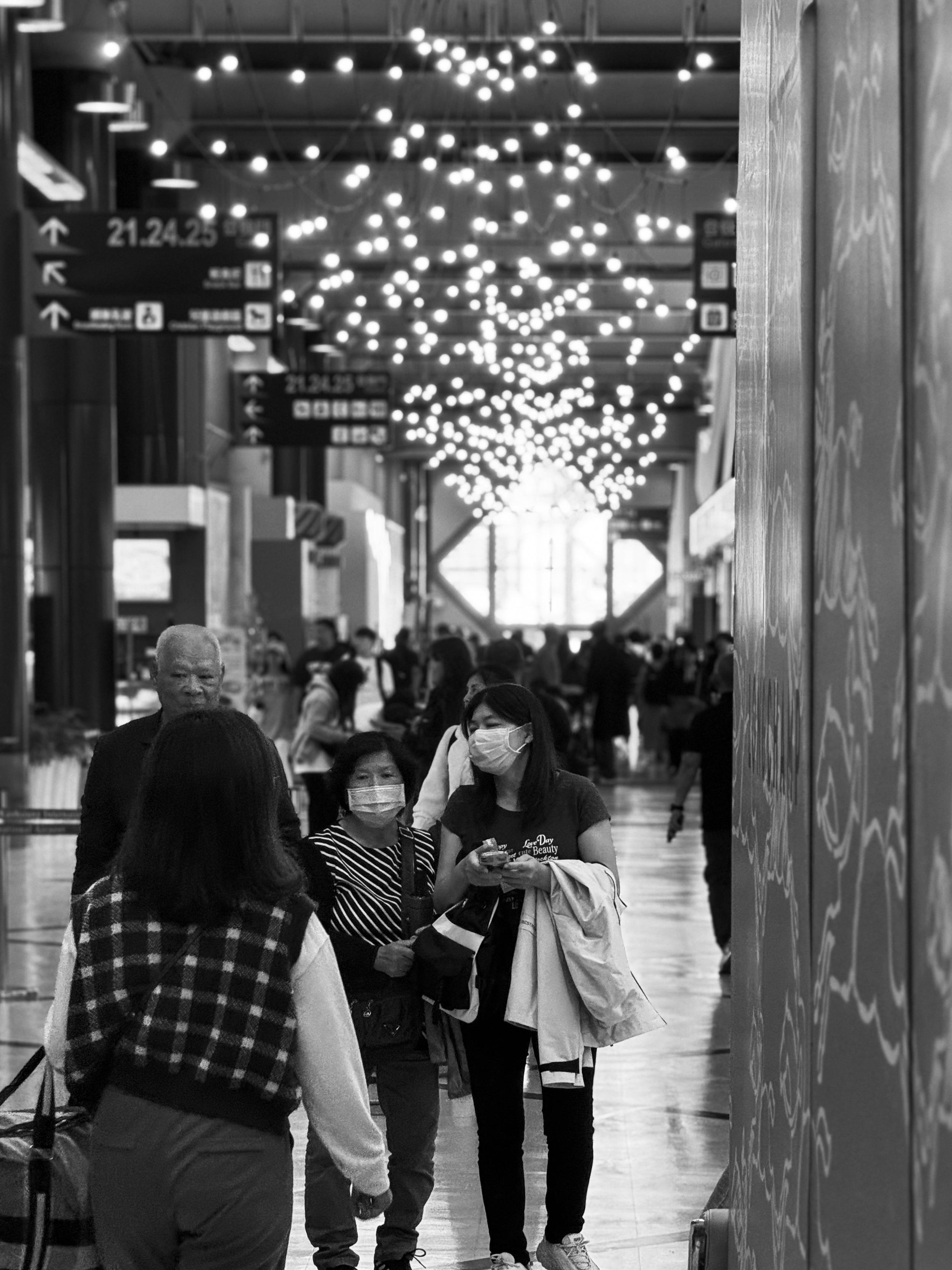 People wearing face masks and walking inside an airport terminal, with decorative lights hanging from the ceiling.