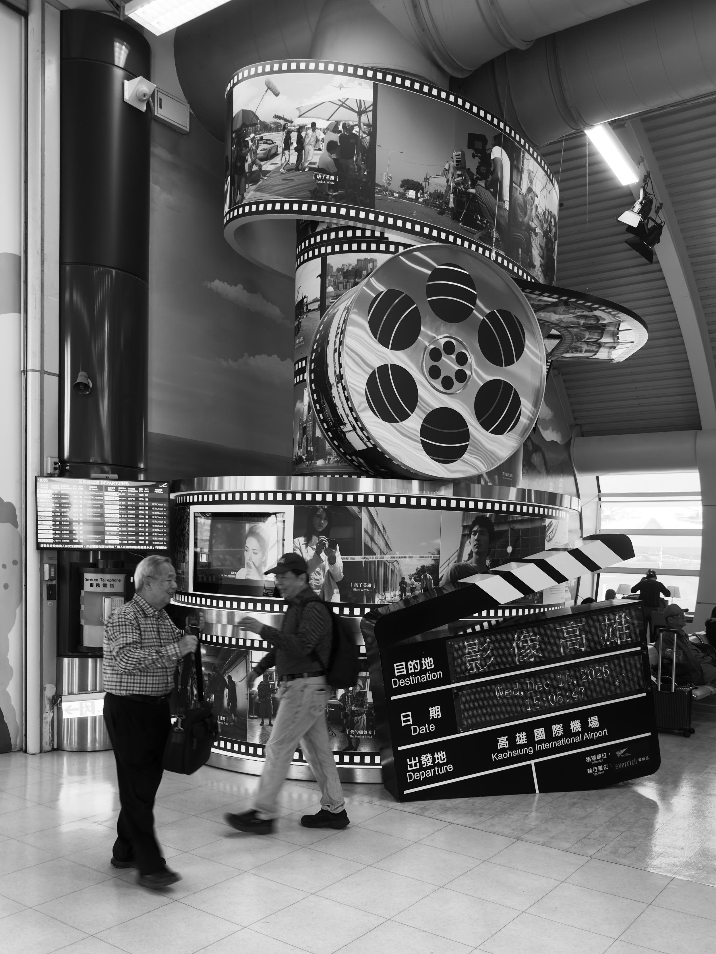 People chatting in front of a movie-themed display at an airport terminal, with flight information for Kaohsiung International Airport.