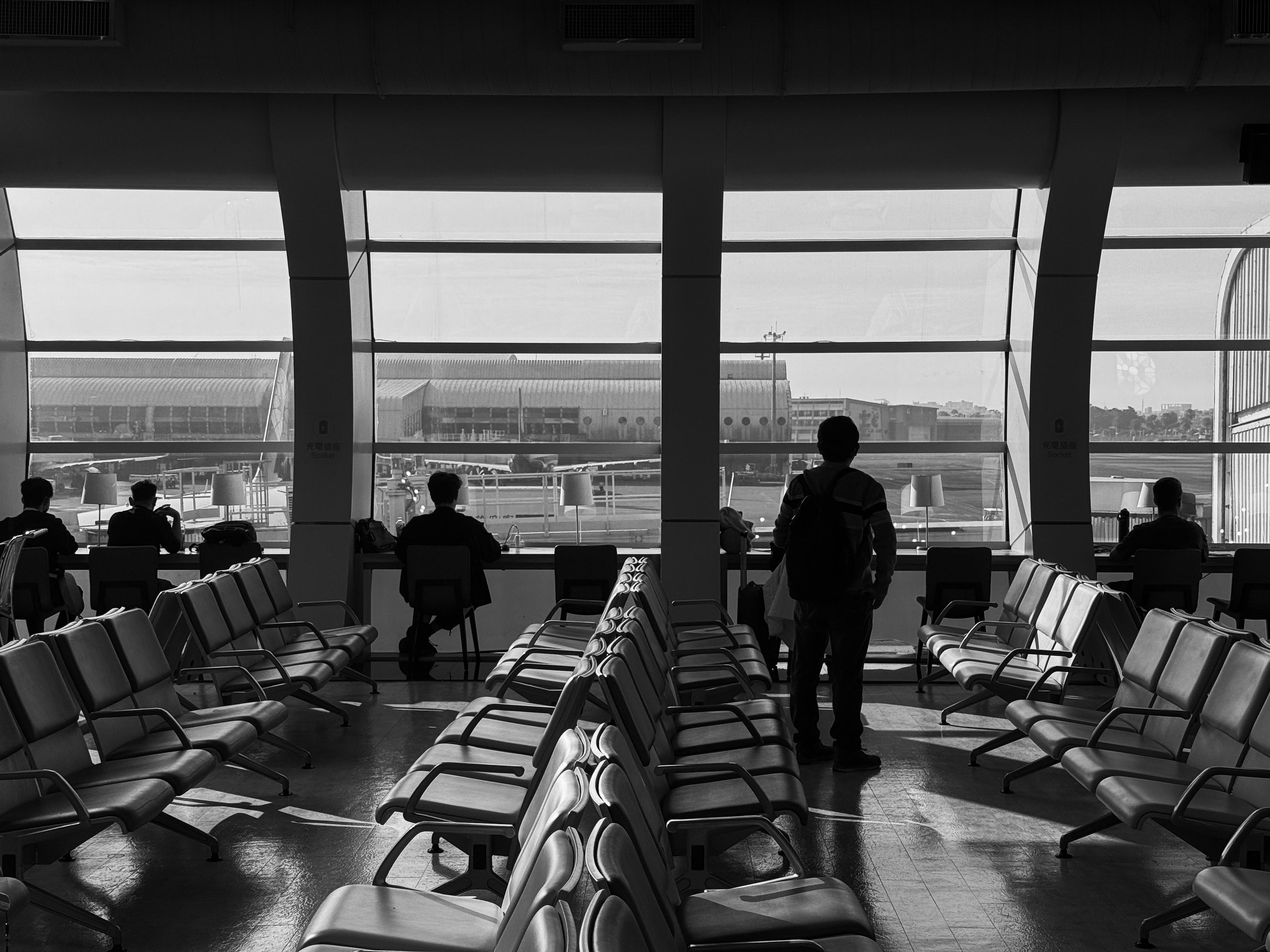 Silhouettes of travelers waiting at an airport terminal indoors, with large windows overlooking the runway and terminal exterior.