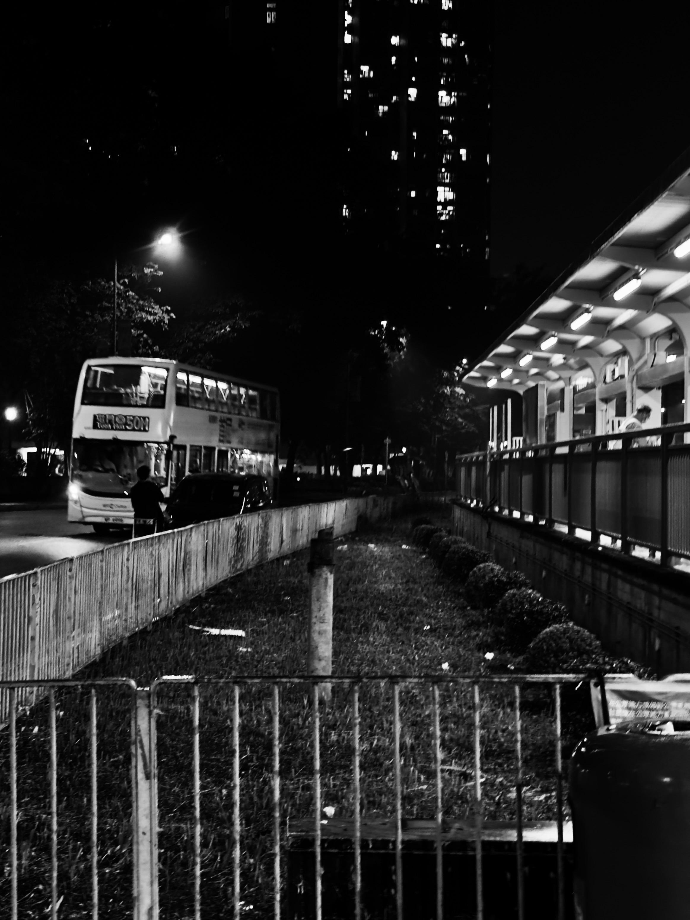 夜晚街道，公交车站有两个人，一个人在等车，背景有高楼和街灯照明。