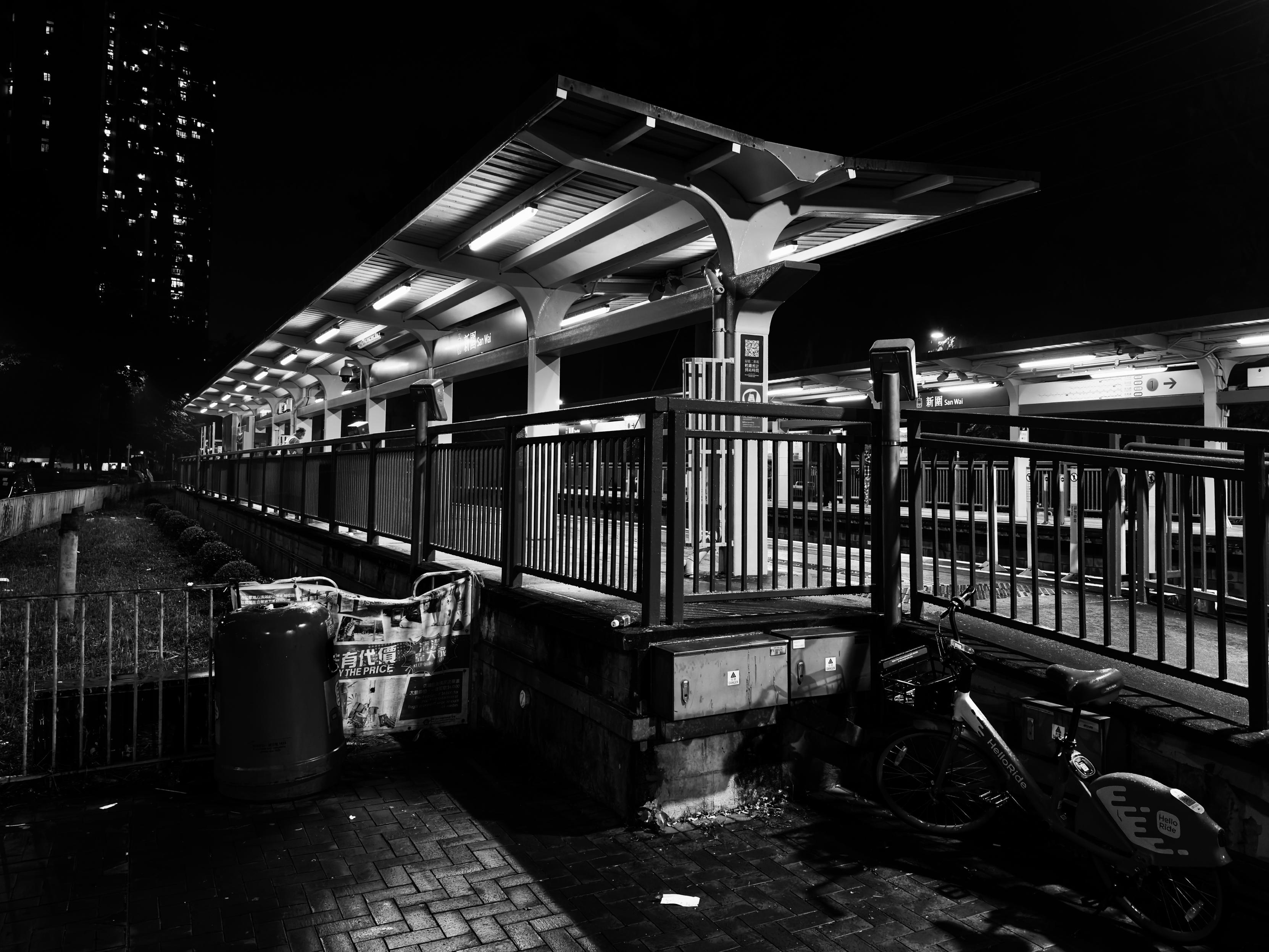 夜晚的巴士站台，黑白色调灯光照明，旁边有自行车和垃圾桶。