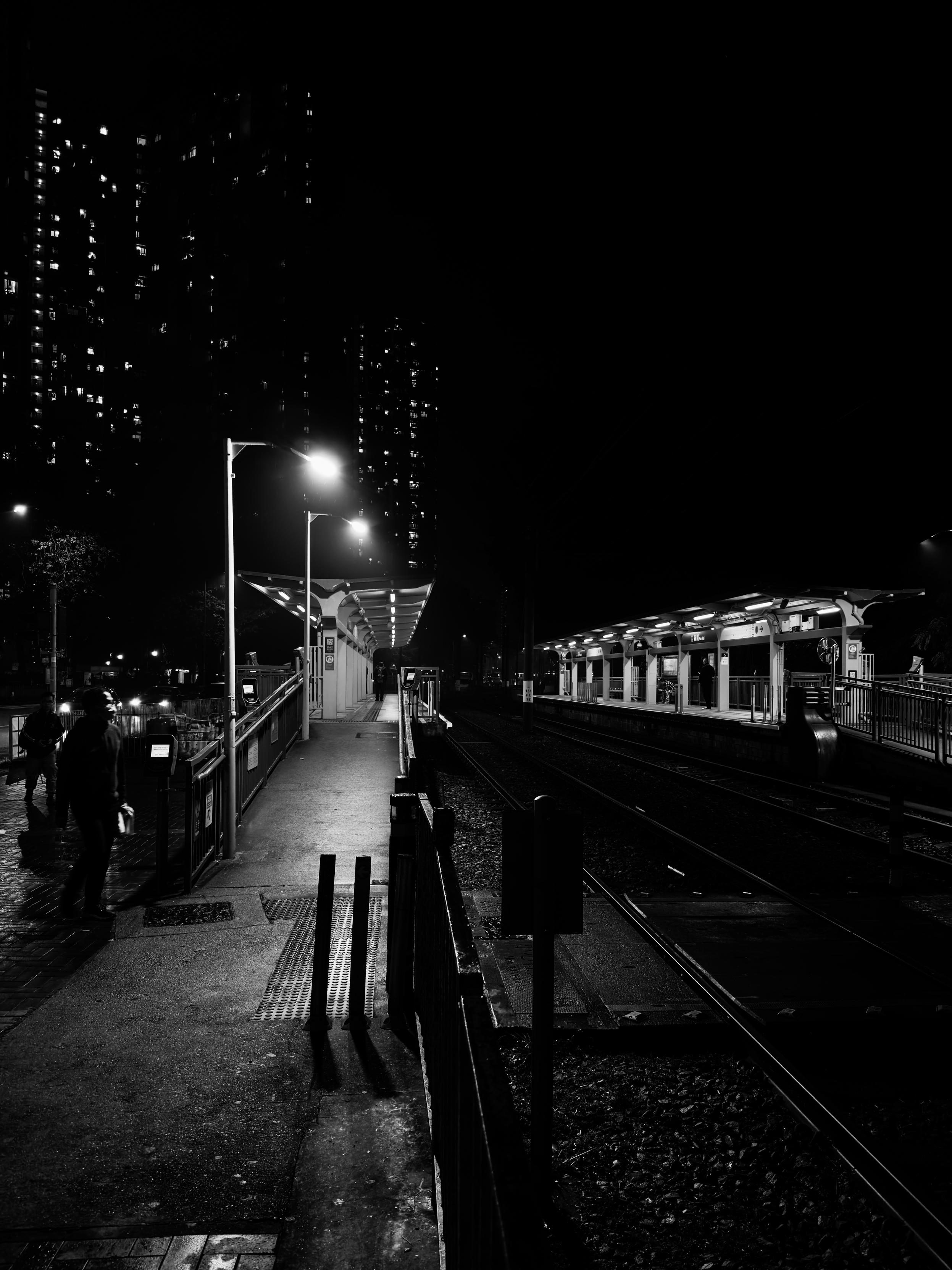 黑白夜晚地铁站，站台上空无一人，几位乘客等待列车，城市高楼在背后点缀灯火。