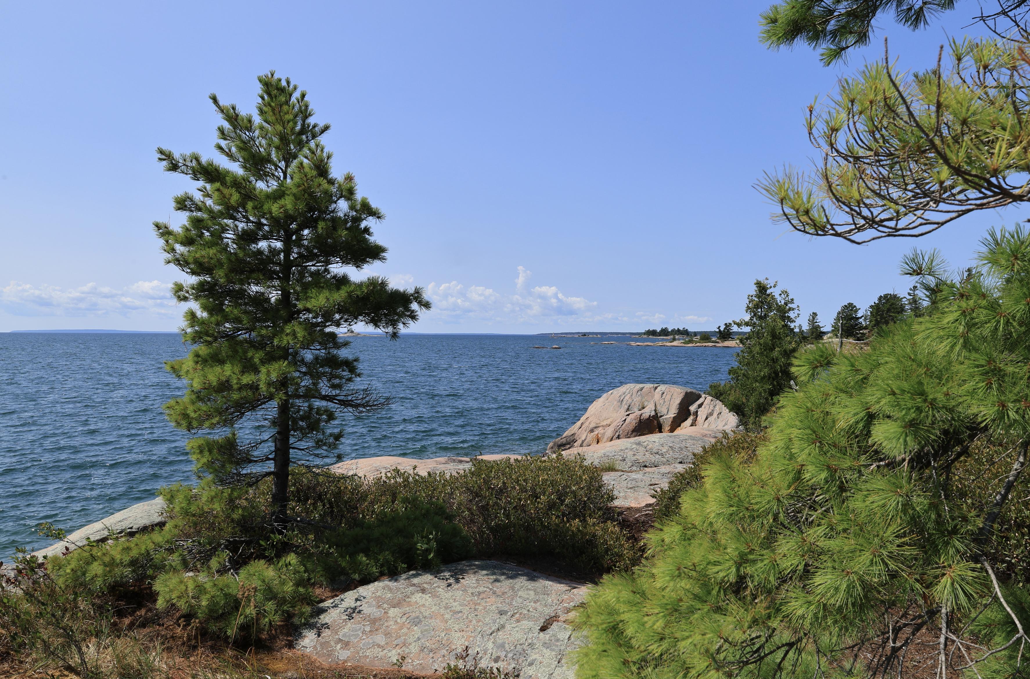This is a landscape photo taken in the summer from a rocky shoreline looking out over a larger lake.  The sky is an interesting shade of blue with only a few white clouds near the horizon. Some coniferous trees can be seen at differing distances. Smaller waves can be seen on the water's surface.