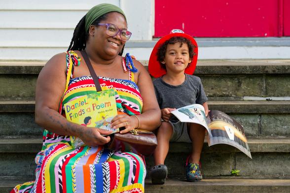 Black woman with multi-colored dress. exposed soldiers and scarf around hair holds book net to a black child wearing a kid's firefighter hat and holding another books on the grey stone steps of a white church with a red door