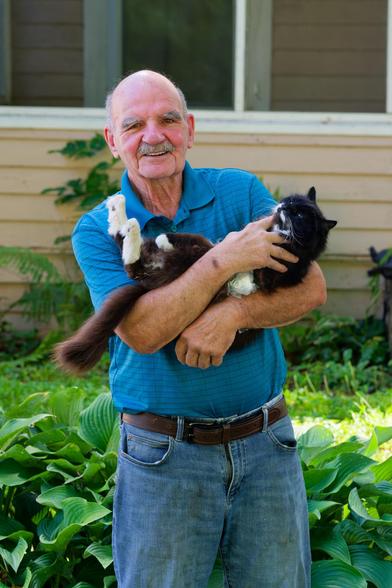 Man in blue shirt looks at me with a slight smile holding a mostly dark cat with white spots on its feet and elsewhere in front of hostas, a lawn and a light yellow house