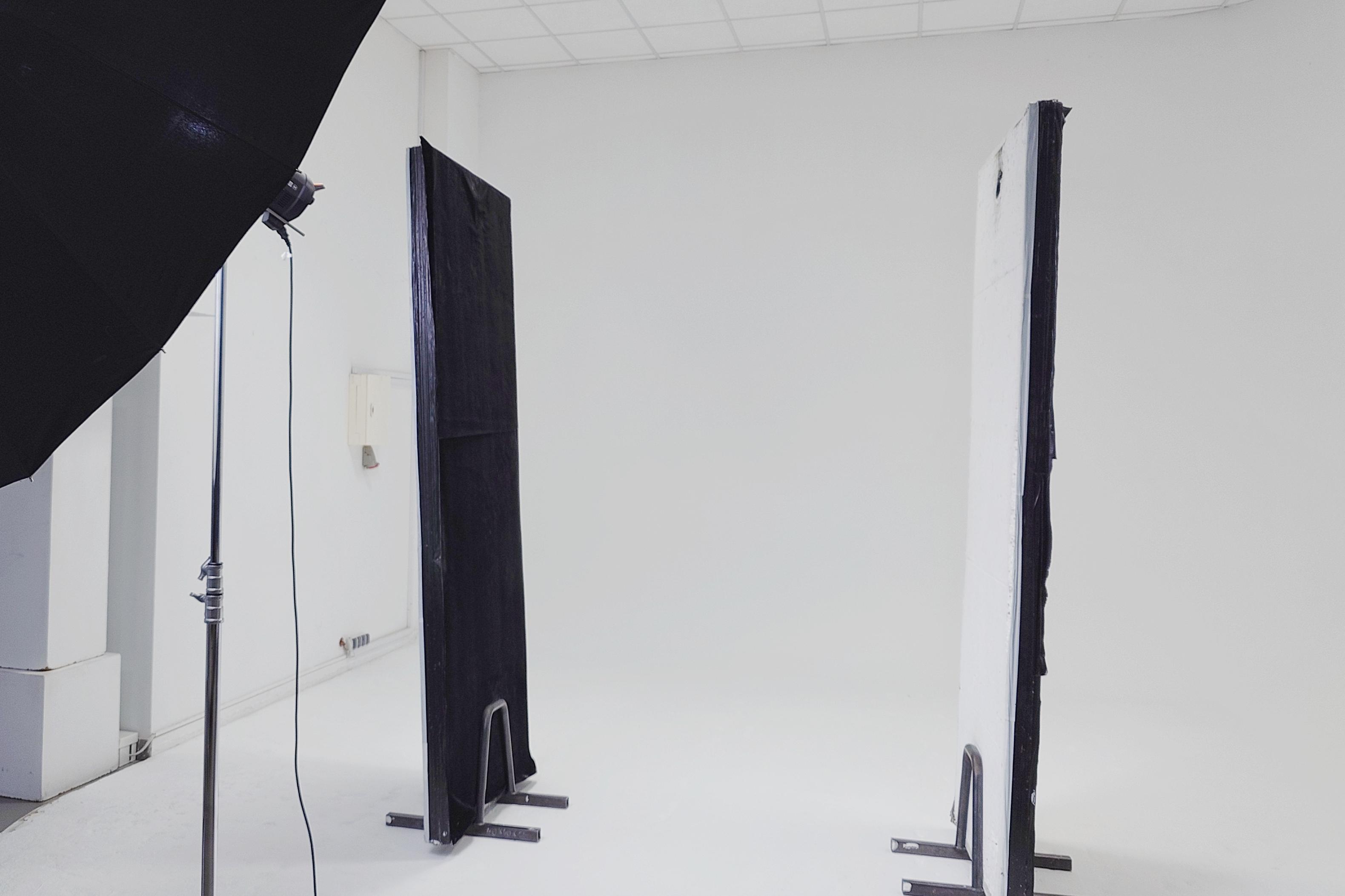 Szerokie ujęcie profesjonalnego studia fotograficznego - Studio Stawki, z białą cykloramą. Po lewej stronie widać fragment dużej, czarnej octy (modyfikatora światła) zamontowanej na wysokim statywie oświetleniowym. W centralnej części stoją dwa wolnostojące, prostokątne panele (V-flats). Lewy panel jest cały czarny, a prawy panel ma białą powierzchnię skierowaną w stronę kamery. Oba panele stoją na niskich, metalowych statywach. Tło stanowią gładkie, białe ściany i podłoga przechodząca w cykloramę. W głębi na ścianie widać małą, białą skrzynkę przyłączeniową. Pomieszczenie ma sufit kasetonowy. Przestrzeń jest pusta, gotowa do sesji zdjęciowej.