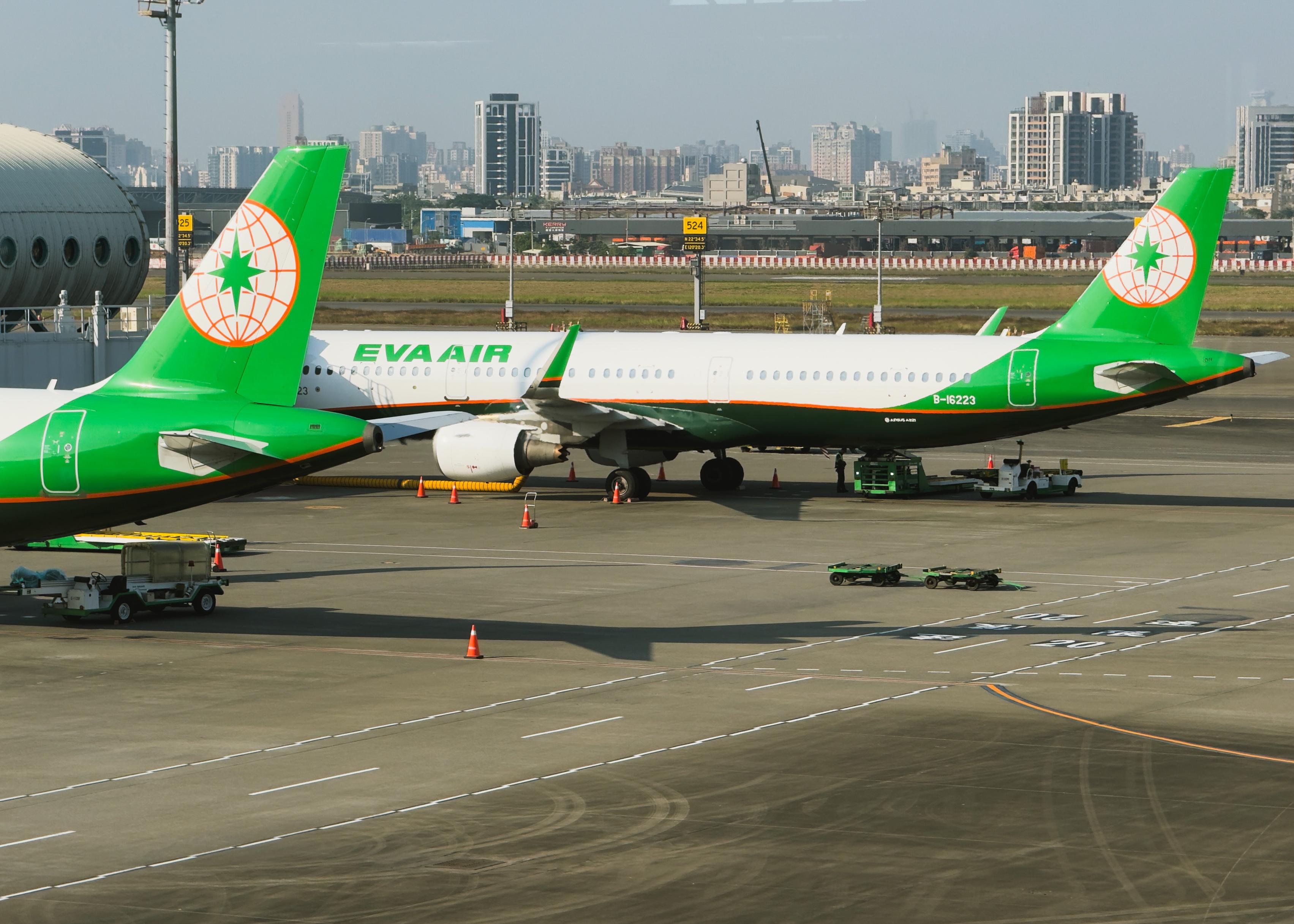两架绿色与白色的EVA Air飞机在机场停靠，背景是城市天际线。