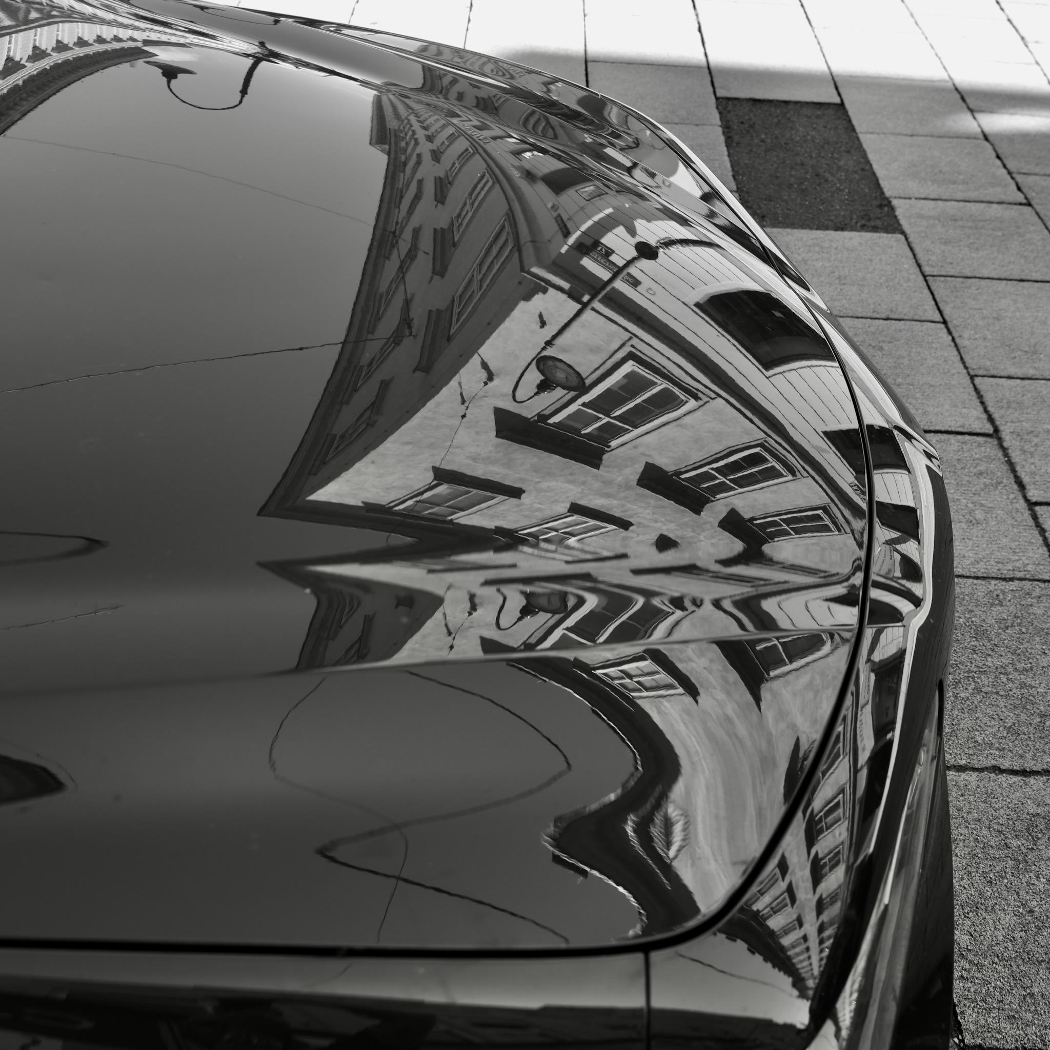 Picture of a car hood in a city. The surrounding buildings are reflected on the shiny hood and are distorted by the form of the metal. The houses are curved and formed in unusual ways. 
Background are plaster stones. Photo is black and white.