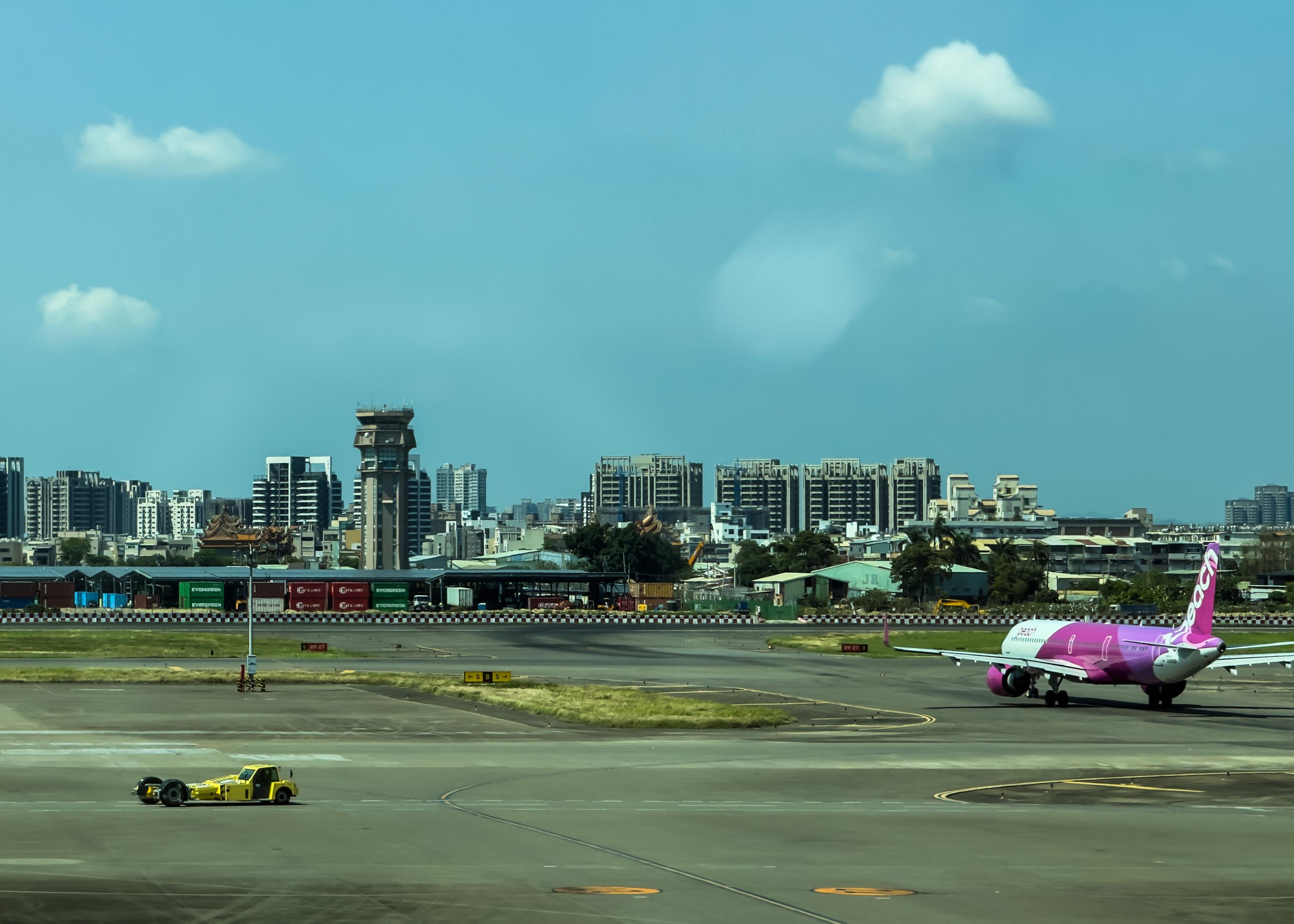 An airplane on the runway with a city skyline in the background, under a partly cloudy blue sky.