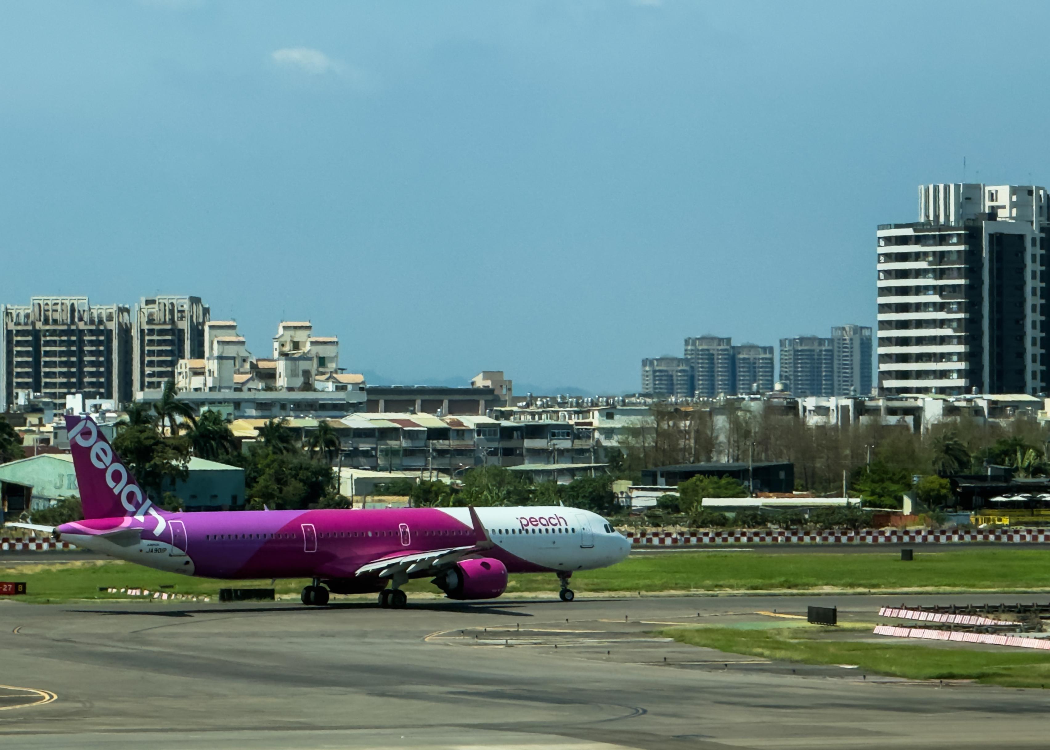 A pink and white Peach airline airplane on the runway with a city skyline of tall buildings in the background.