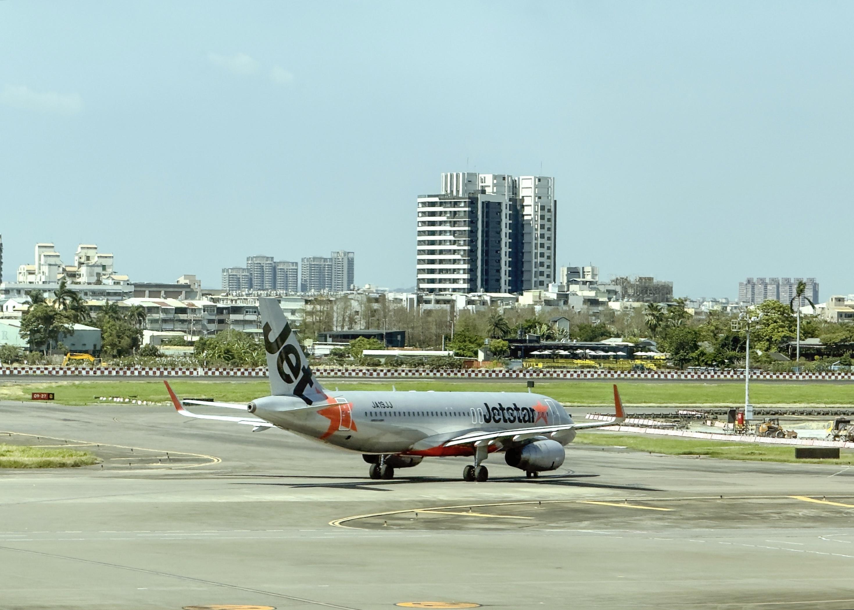 一架Jetstar飞机在机场跑道上准备起飞或降落，背景是城市建筑和蓝天。