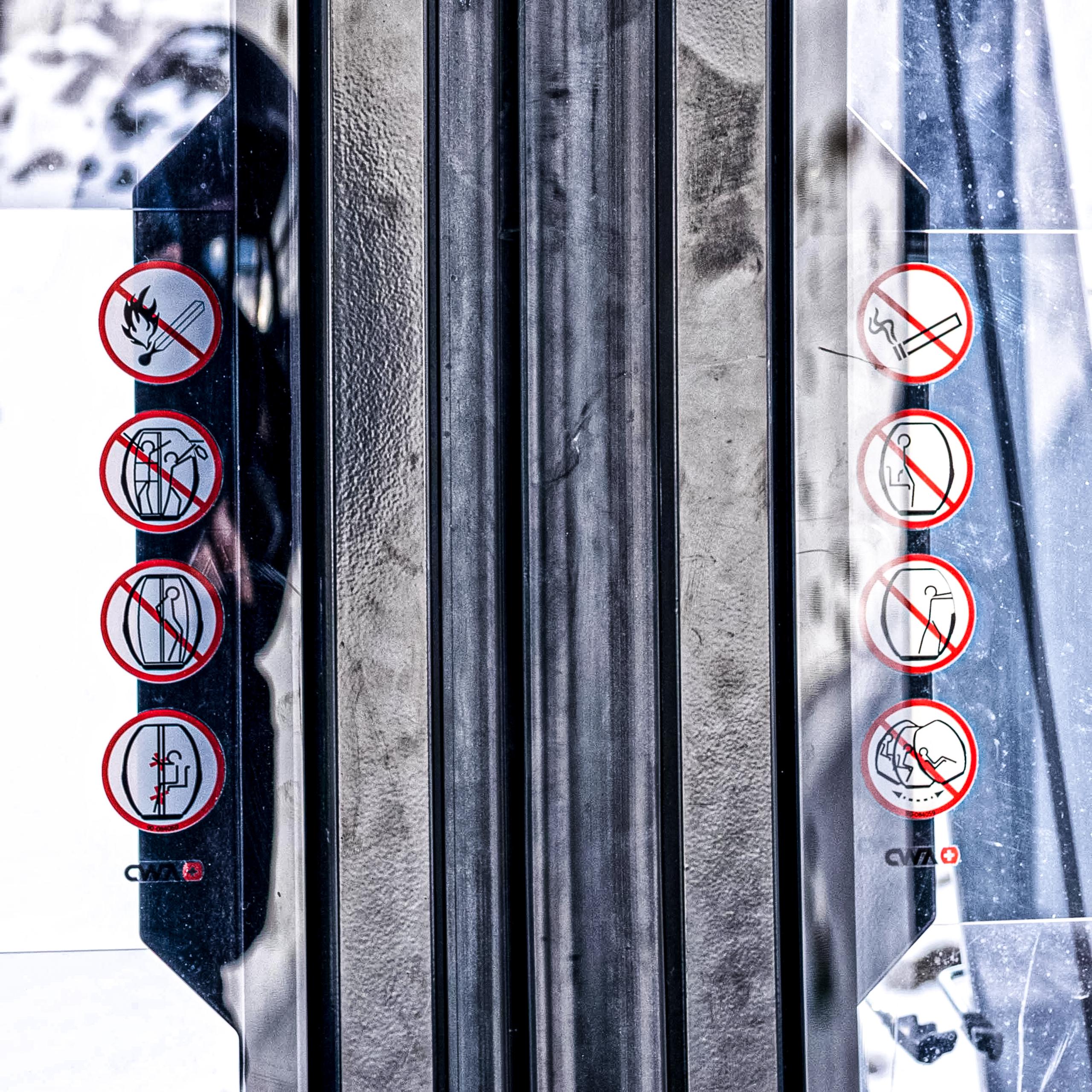 Nahaufnahme der automatischen Schiebetüren einer Seilbahnkabine (Hersteller CWA). In der Mitte verlaufen die vertikalen, metallischen Türleisten und Gummidichtungen. Auf den Glasscheiben links und rechts sind spiegelbildlich angeordnete, runde Verbotsschilder mit roten Piktogrammen auf weissem Grund angebracht, die Sicherheitsregeln wie Rauchverbot, offenes Feuer oder Hinauslehnen untersagen. In den Scheiben spiegeln sich das Umfeld und eine verschneite Kulisse.
