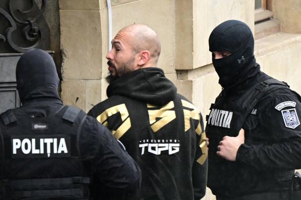 Man in black hoodie with gold and white symbols and shaved head takes perp walk between two cops wearing dark clothes that say POLITIA.