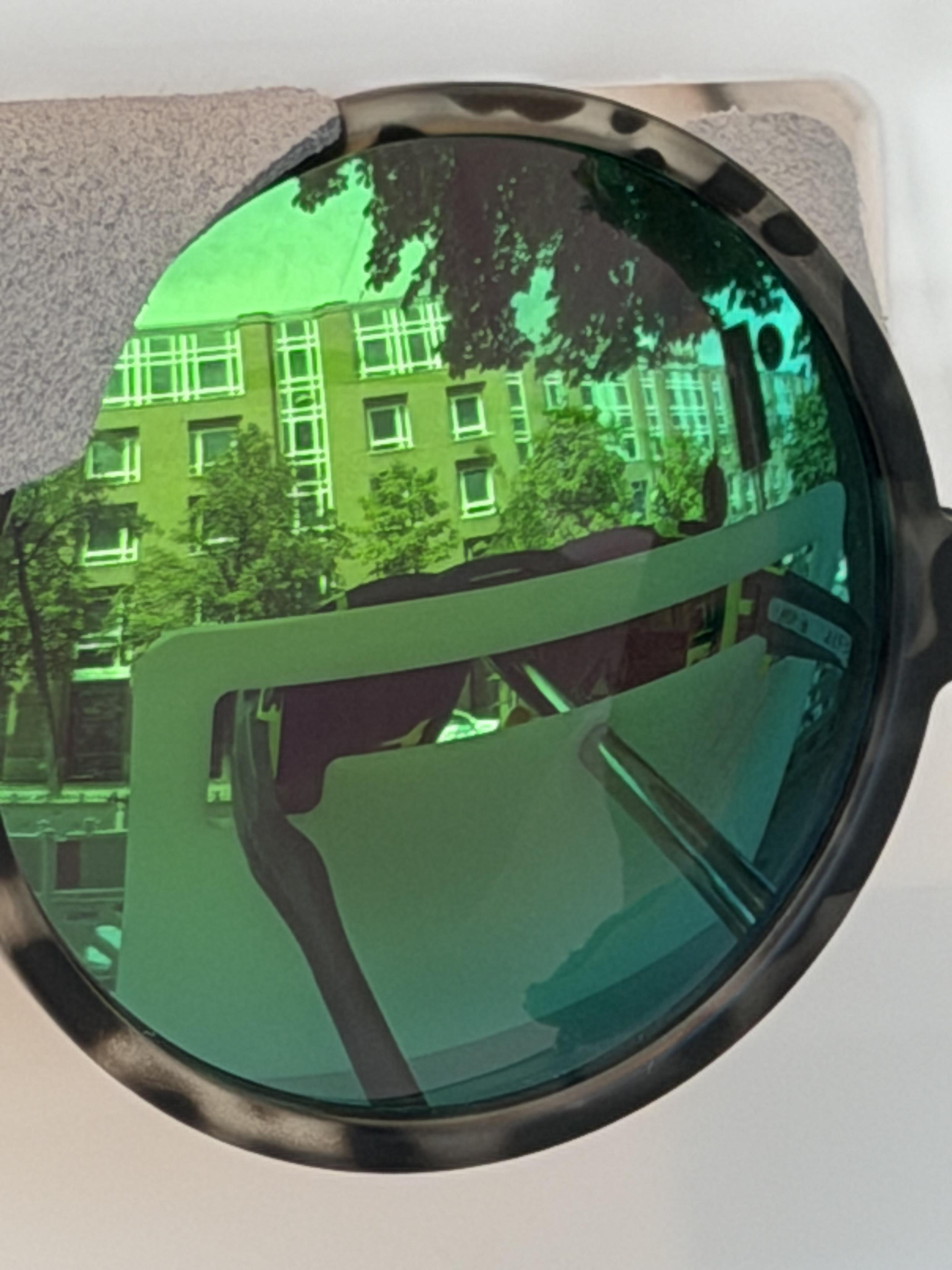 Close-up of a large round green-tinted sunglass lens reflecting an urban street with apartment buildings, trees and part of the surrounding structure. The reflection fills almost the entire lens.