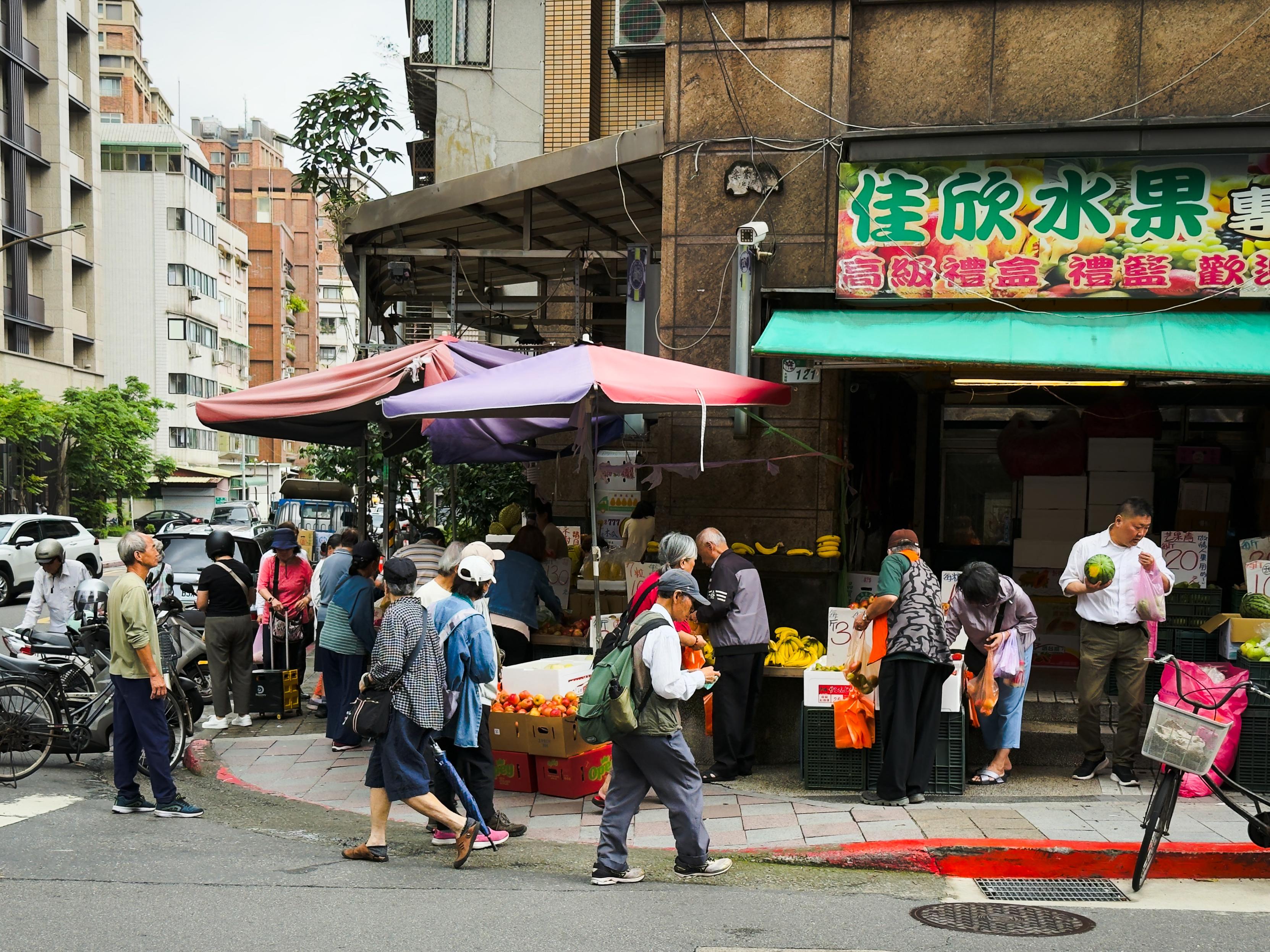 繁忙的街边水果摊，许多老人购买水果，背景是多层建筑和悬挂的彩色广告牌。