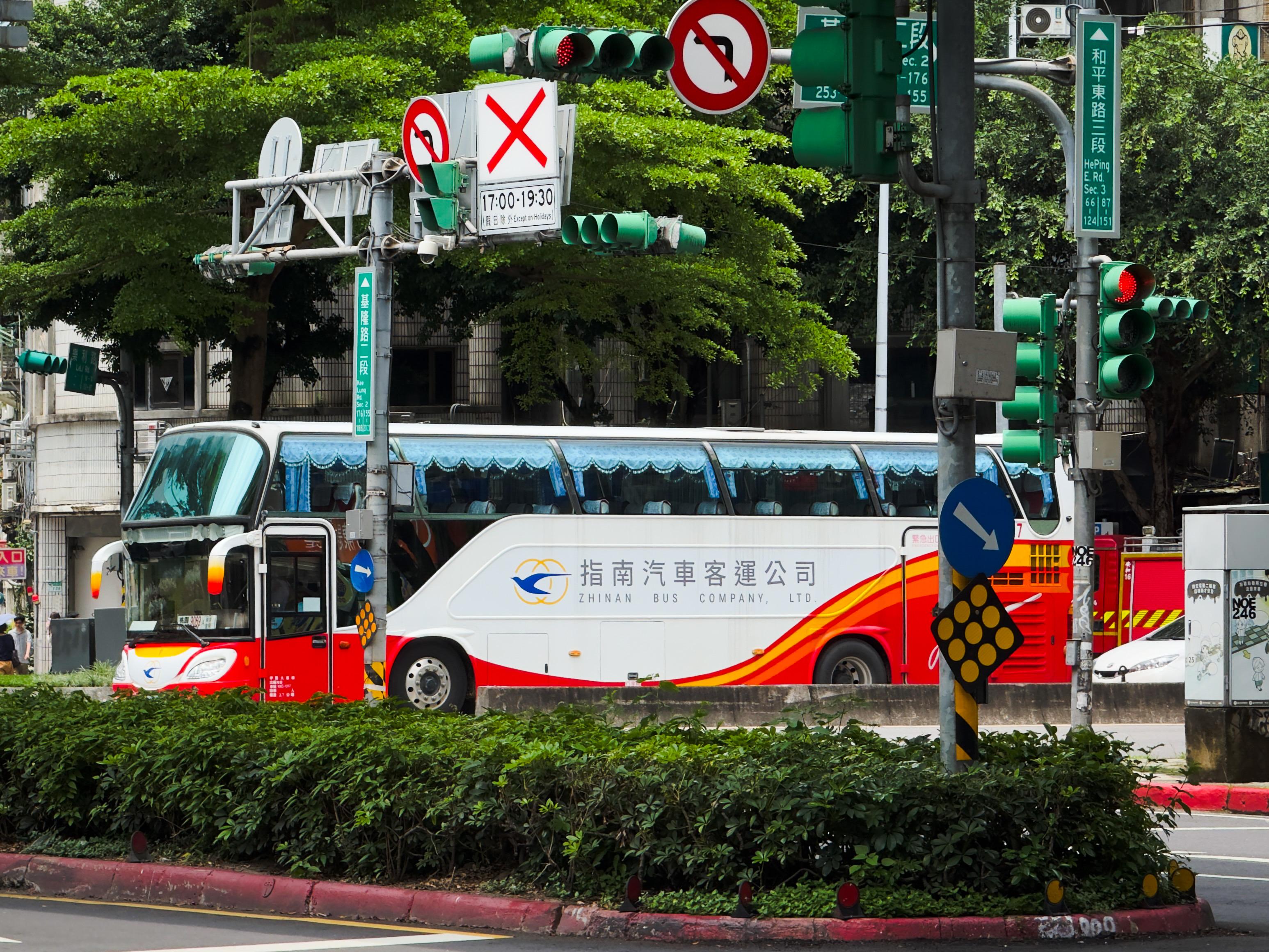 一辆白色和红色的双层巴士在城市街道上行驶，周围有交通信号灯和绿树。
