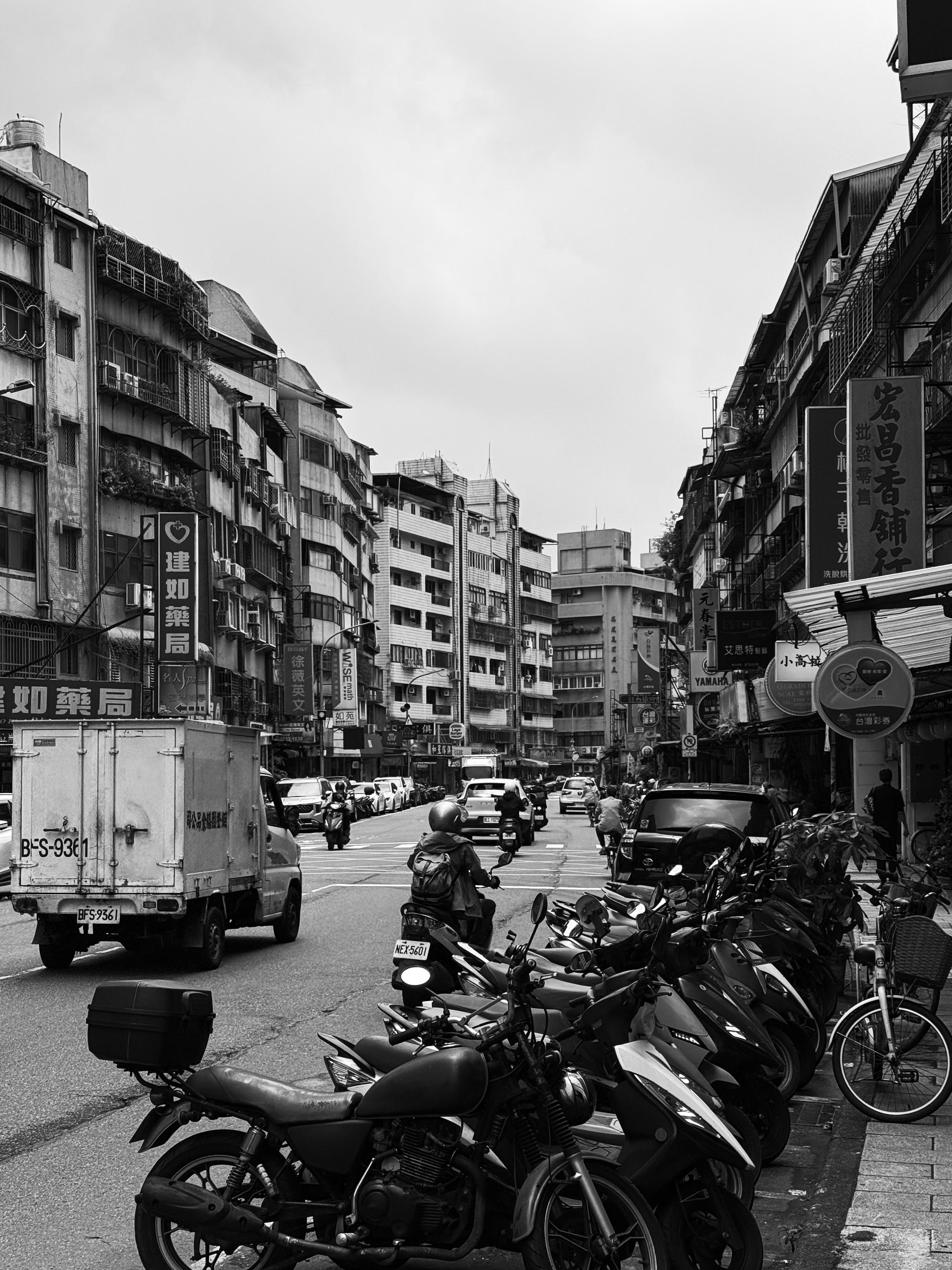 繁忙街道上的摩托车和汽车，左侧有一辆货车，背景是多层建筑和商店招牌。