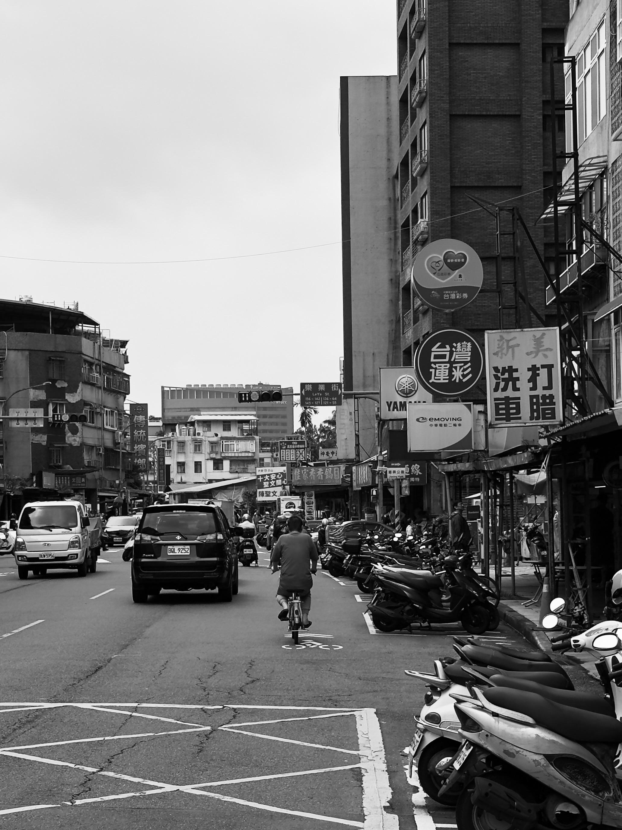 繁忙街道，行人騎自行車，停放許多電動車與摩托車，背後有多層建築和商店招牌。