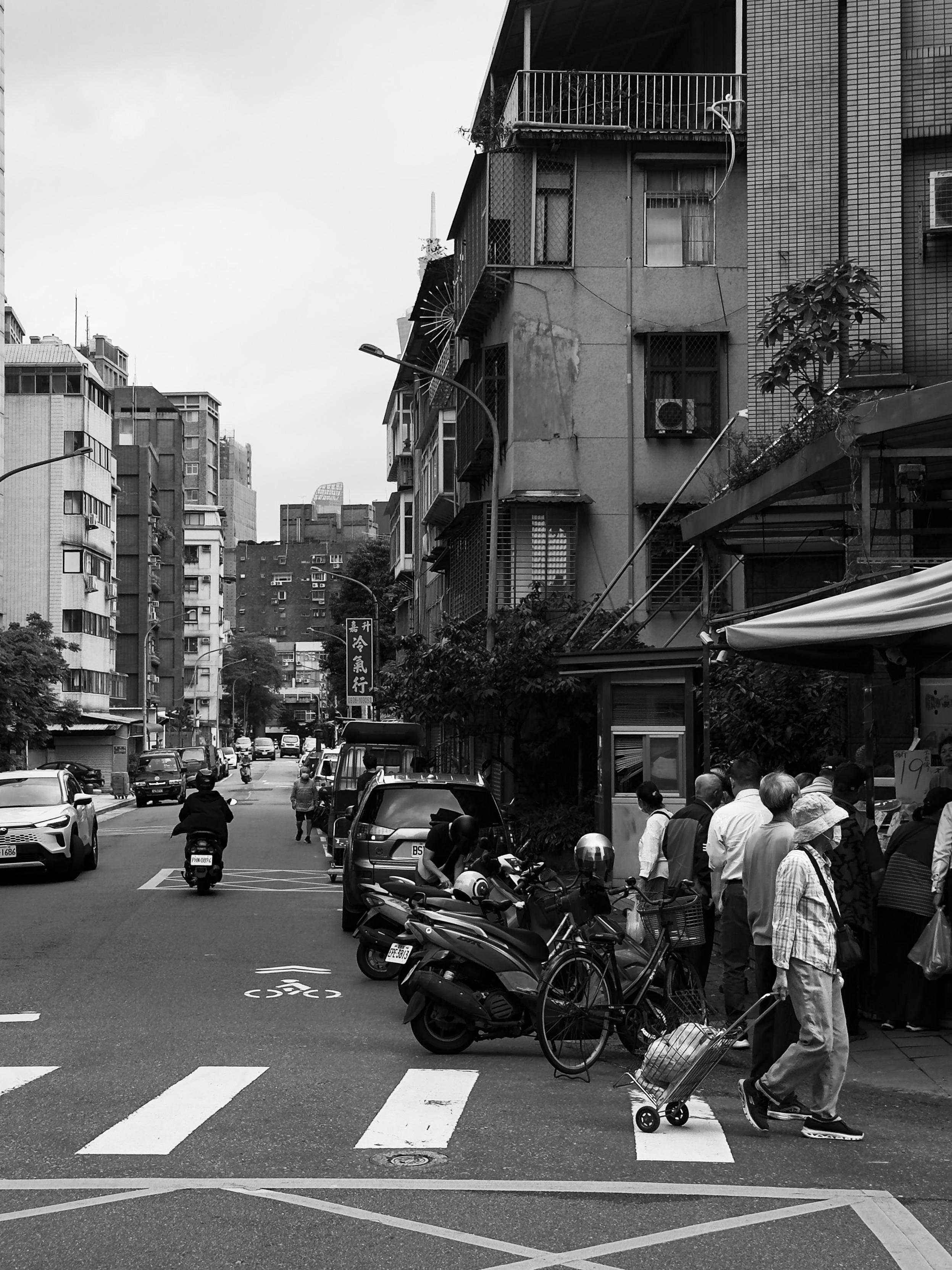 街道上有人排队买东西，街边停放多辆摩托车和自行车，背景是多层老旧建筑。
