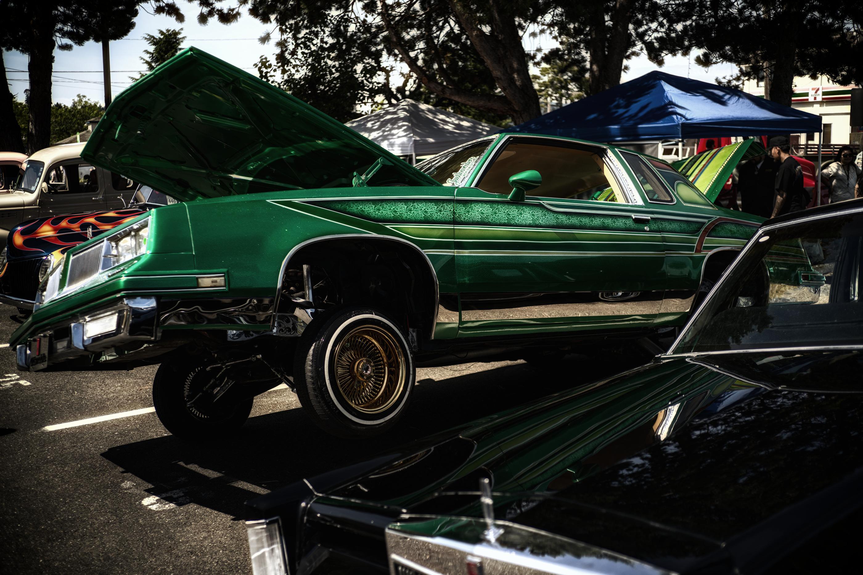 Color photo in portrait format of a dark green custom low rider Oldsmobile Cutlass (Supreme or 442) from the late 1970's or 1980's.

The low rider fills the entire center of the photo. Custom hydraulics raise the driver side tire off the ground at a 30 degree angle, about 25-35 cm off the ground. Custom gold spoked wheel rims shine brightly, as do the custom chrome colored suspension and hydraulics visible in the wheel well and underside. Horizontal pin striping outlined in white runs from rear trunk to engine bay in five sections, with the topmost section decorated in a flower pattern. A sticker that reads "lowriders worldwide" is attached to the bottom corner of the driver side windshield. Hood and rear trunk are open for display. 

Another classic black Oldsmobile from the 1960's or 1970's is in the bottom left quadrant of the photo with half of it's front windshield and engine hood visible.

To the left of the green Cutlass are three more partially visible classic cars. Behind the 