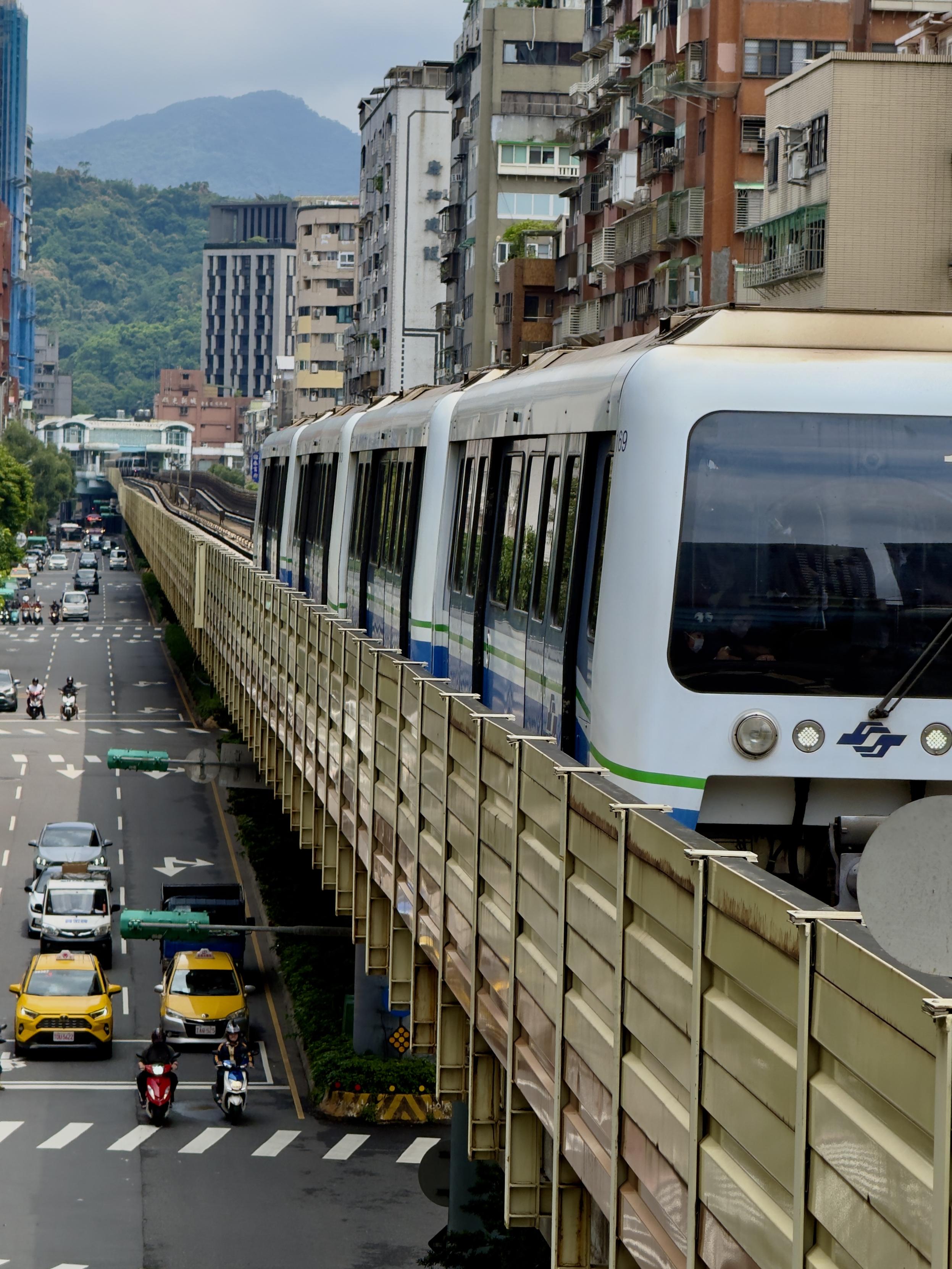 这是城市街道上的高架轨道列车，周围是繁忙的交通和多层建筑，背景有山峦。