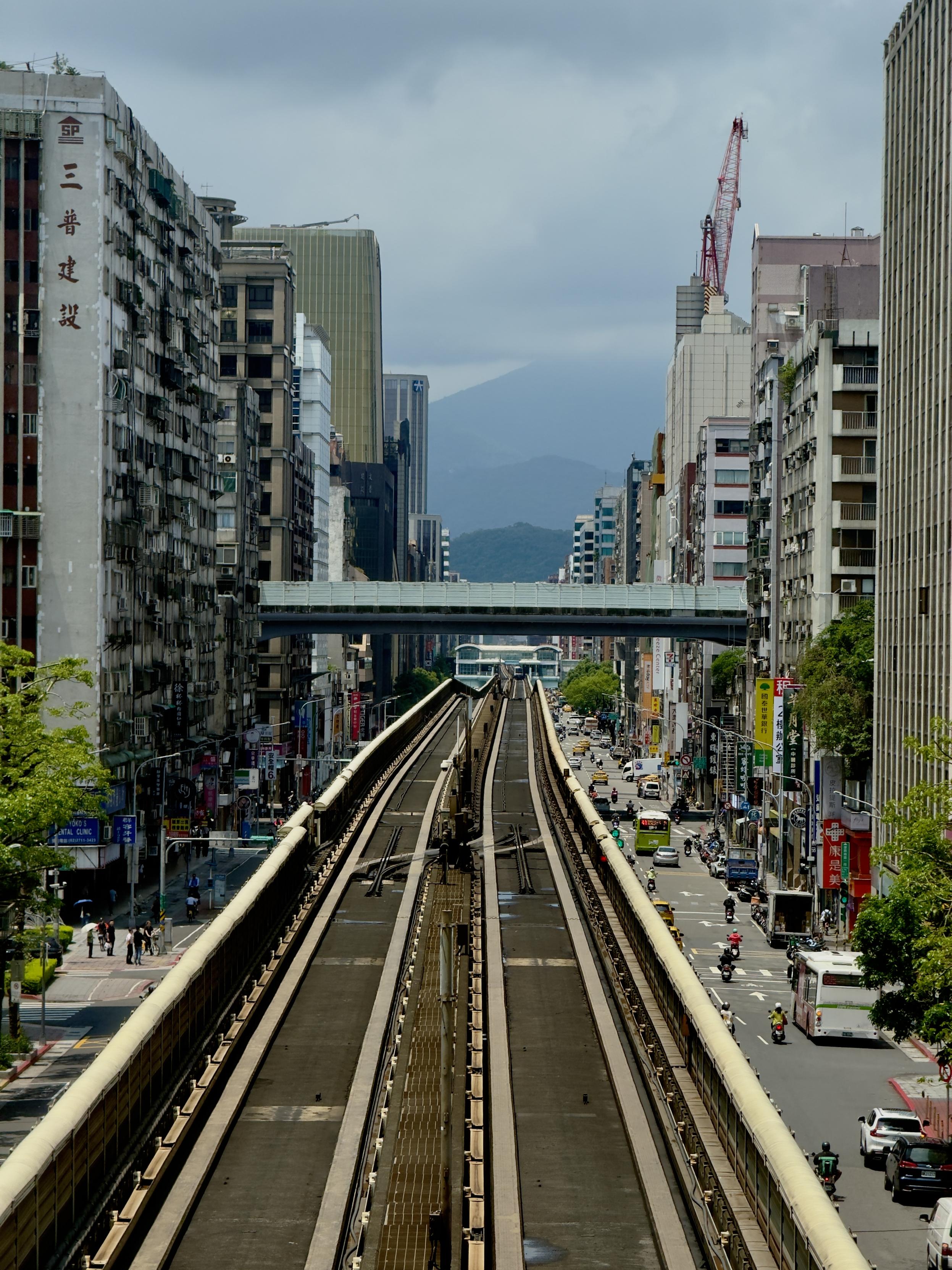 繁忙的城市街道和高架鐵路穿越高樓林立的街區，遠處可見山脈與部分山頂。