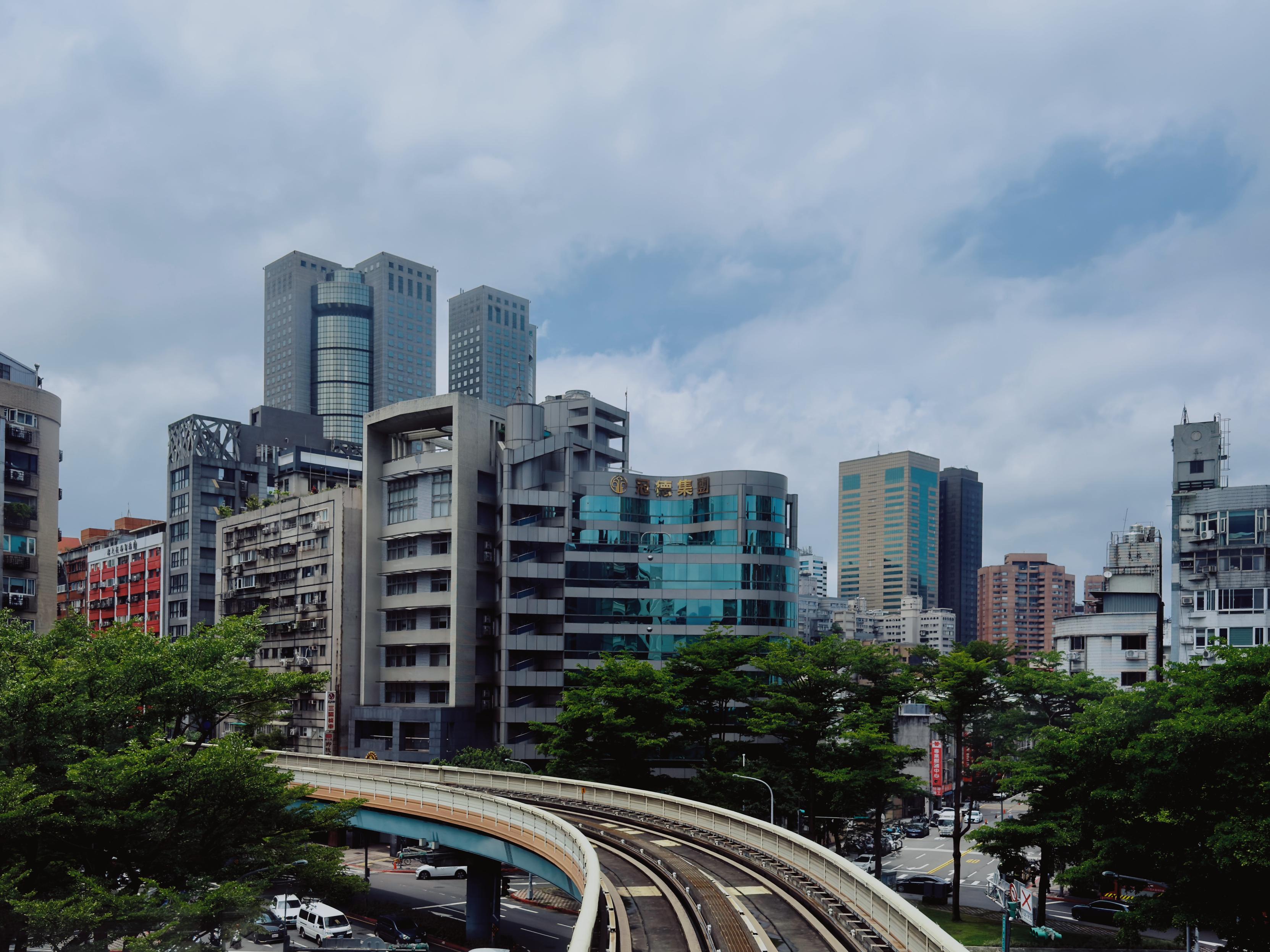繁忙都市景观，摩天大楼高耸入云，城市道路环绕，绿树点缀，天空布满云朵。