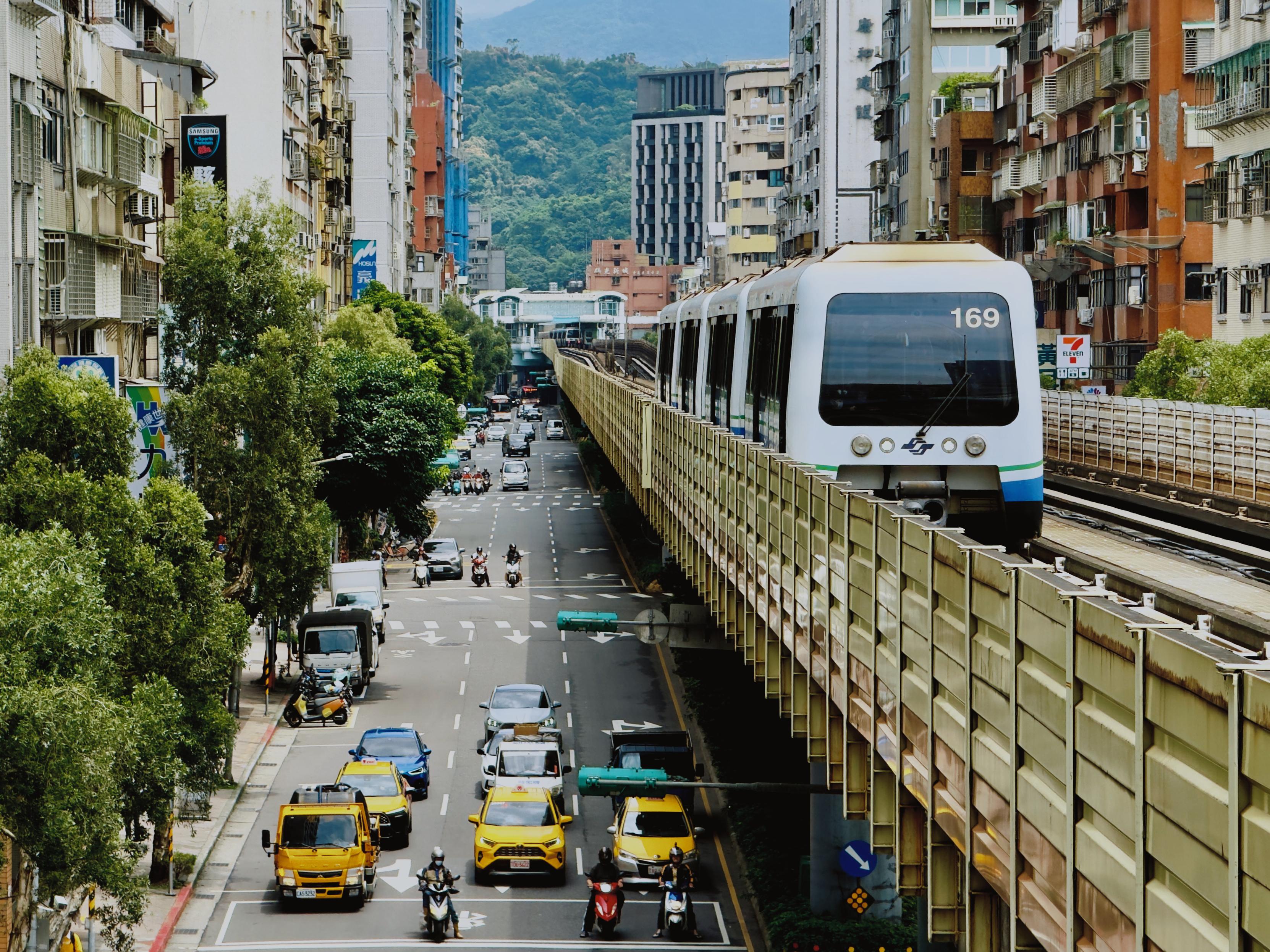 一辆白色地铁列车在繁忙城市道路上行驶，周围有车辆和摩托车，背景是高楼和山峦。