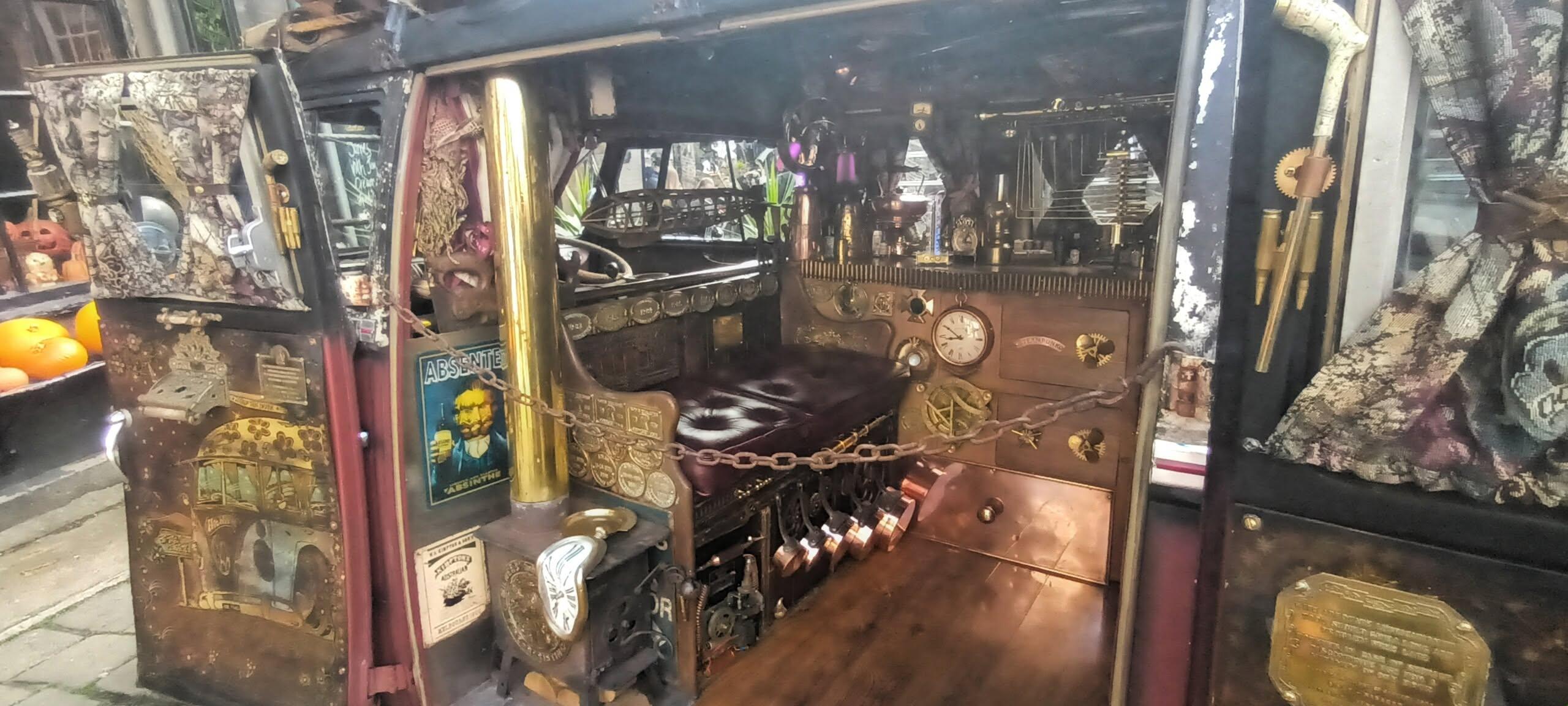 Photo of the interior of a campervan at a steampunk gathering. The double doors are wide open showing an interior decorated with brass, copper, dark leather and wood. By the door is a cast iron wood burning stove with a brass chimney. On top of that is a Salvador Dali melting clock. Behind the chimney a poster for absinthe. Next to the chimney is a 2 seater leather seat, below which are a set of small copper pans and various cogs and machine parts. The back wall is also arrayed with clocks, cogs and strange looking instruments as well as a large brass kettle and an oil lamp.