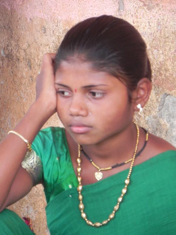 Young woman in a green sari sits against a rough wall, resting her head on her hand and looking downward with a thoughtful expression. She wears gold jewelry and a small red bindi on her forehead.