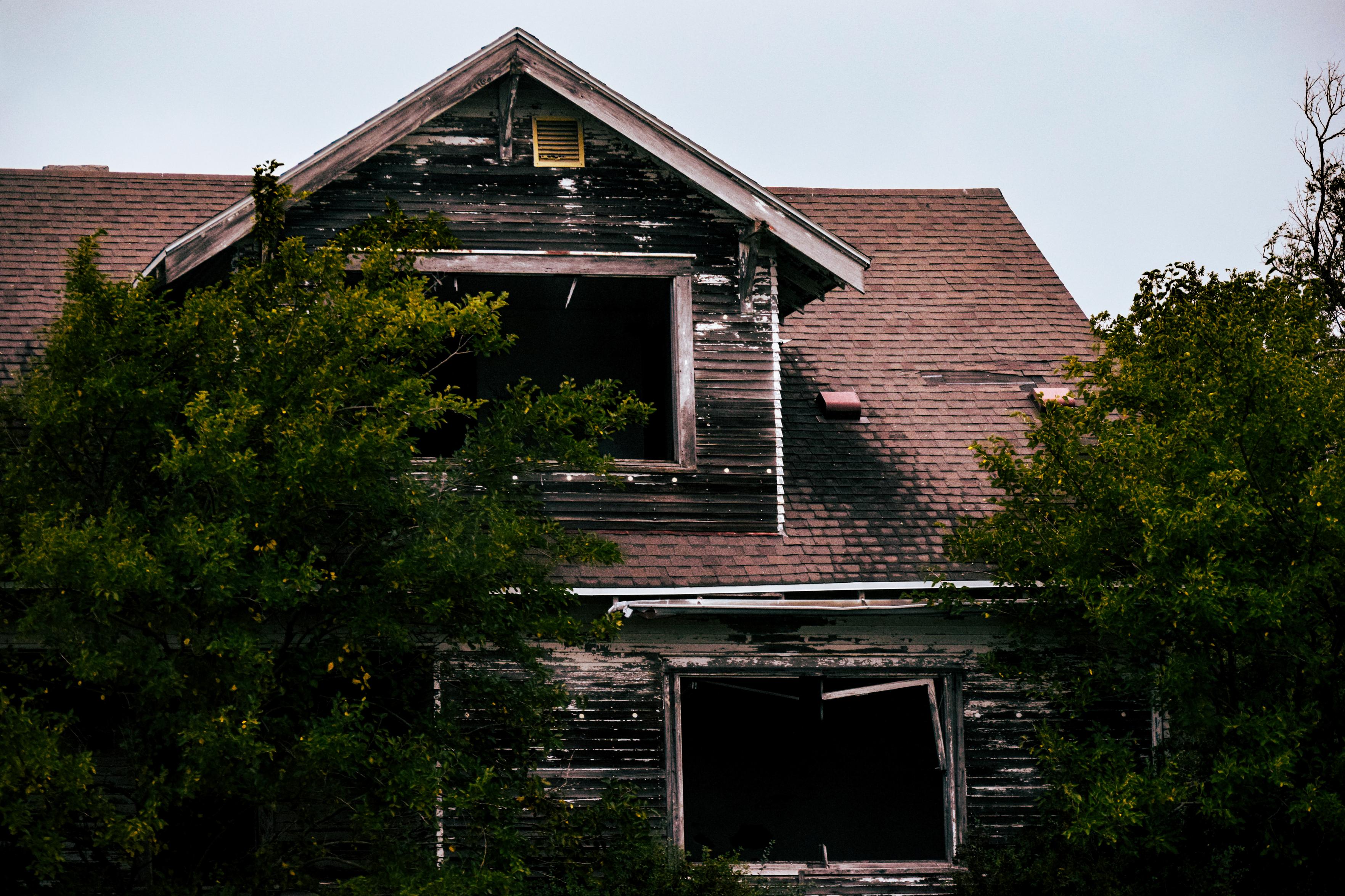 Abandoned wooden house with peeling paint, dark open windows, red-shingled roof, and overgrown trees, creating a haunting, desolate mood.