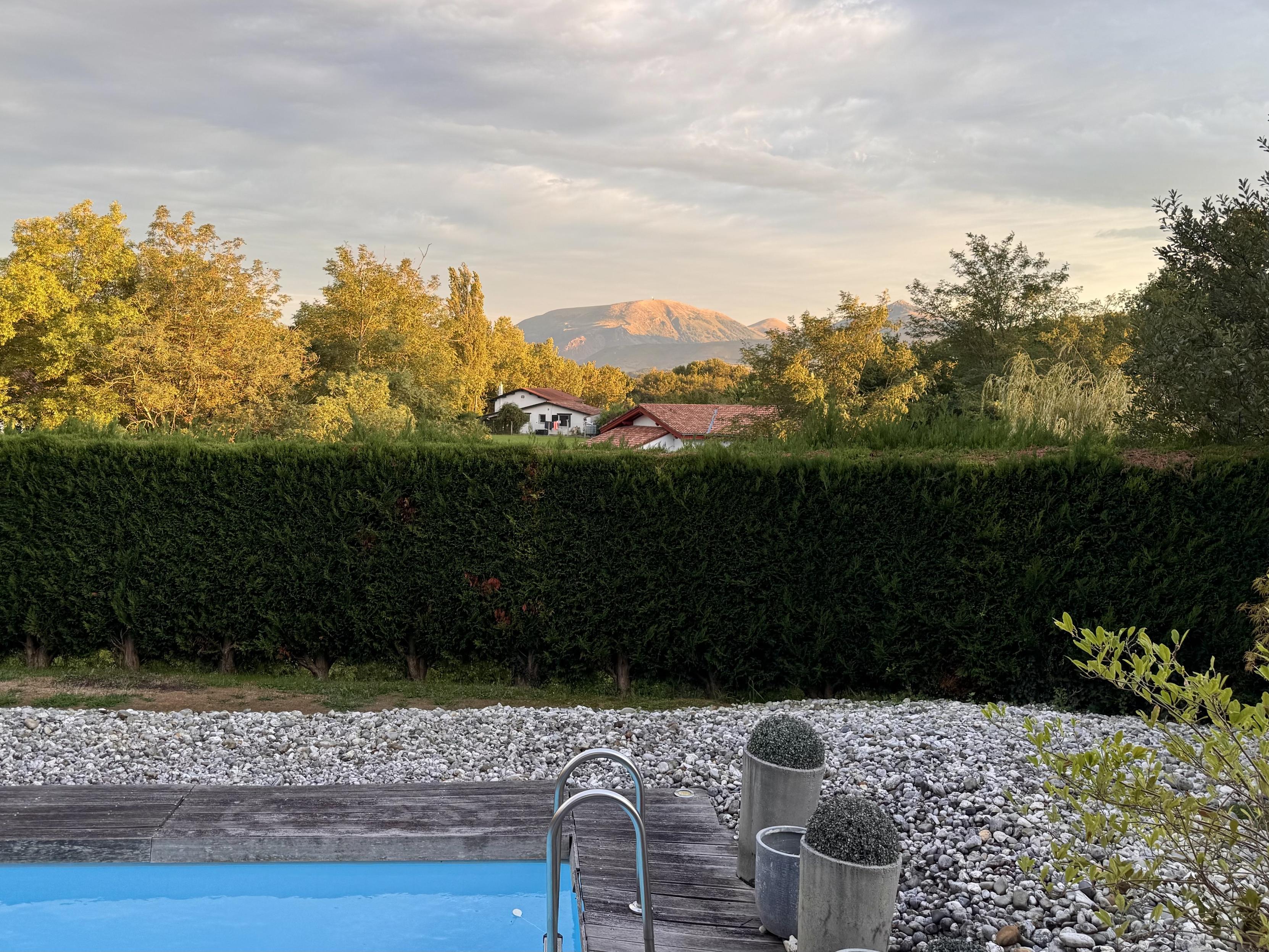 In the background is a round mountain all lit up by evening sun. In the middle ground are two typical basque houses. They are surrounded by tall trees that are also kissed by sunlight. The foreground shows a swimming pools and pebbles behind a neatly trimmed hedge.
