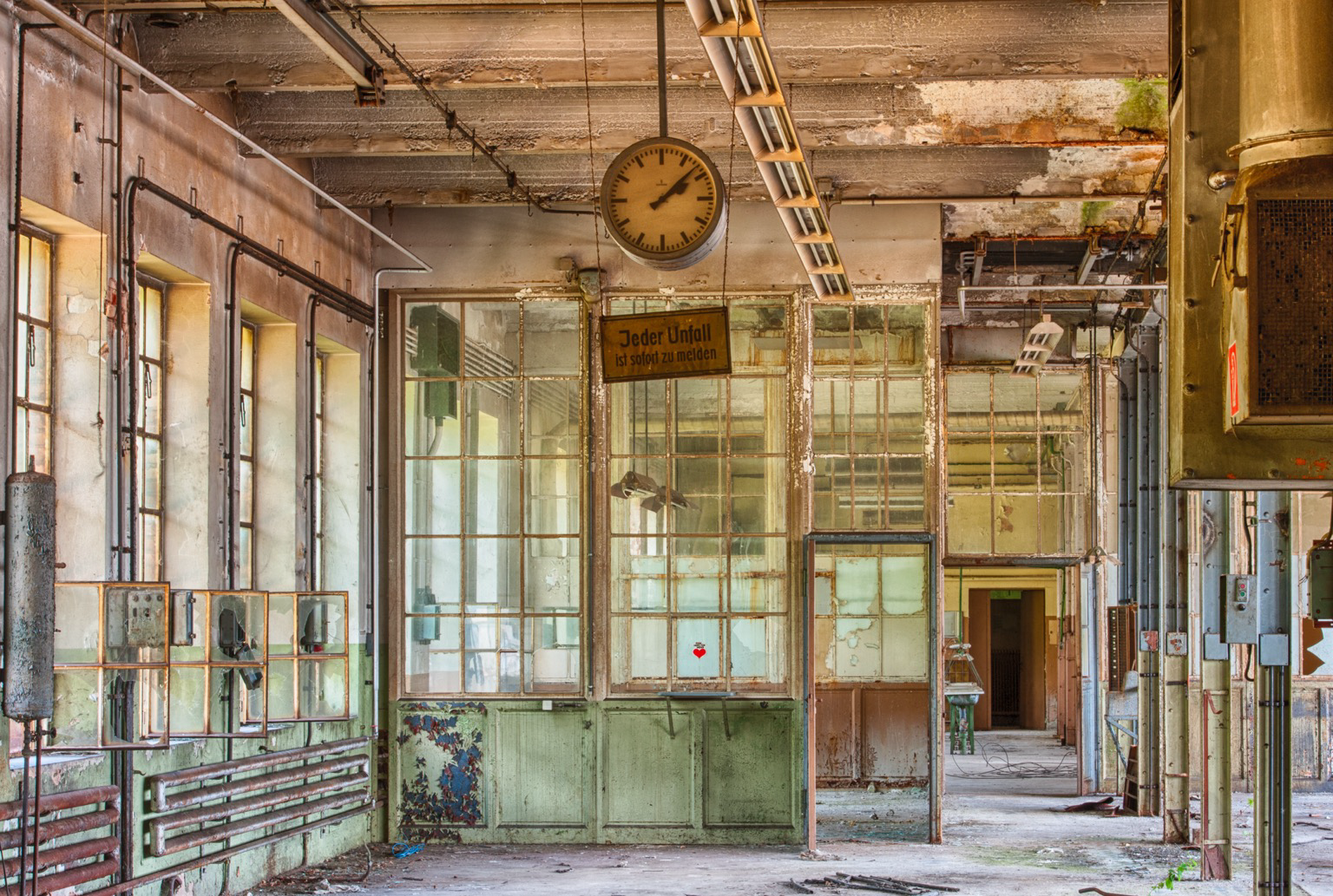 Ein verlassener Industrie- oder Werkstattraum. An der Decke mit freiliegenden Rohren, Kabelschächten und verwittertem Beton hängt zentral eine große, runde Analoguhr. Etwas darunter und weiter hinten ist ein rechteckiges Schild mit der schwarzen Aufschrift "Jeder Unfall ist sofort zu melden" aufgehängt. Die linke Seite und die Mitte des Raumes werden von großflächigen Sprossenfenstern eingenommen. Der untere Teil dieser Fensterfronten ist mit grüner Farbe gestrichen, die stark abblättert. Auf der rechten Seite befinden sich Stützpfeiler und ein massiver, quaderförmiger Metallkasten. Der Fußboden ist aus Beton und mit Schmutz sowie losen Trümmerteilen bedeckt. Im Hintergrund führt ein offener Türrahmen in einen weiteren, tiefer gelegenen Bereich. An abandoned industrial or workshop room. A large, round analog clock hangs centrally from the ceiling, which features exposed pipes, cable trays, and weathered concrete. Slightly below and further back, a rectangular sign is suspended, bearing
