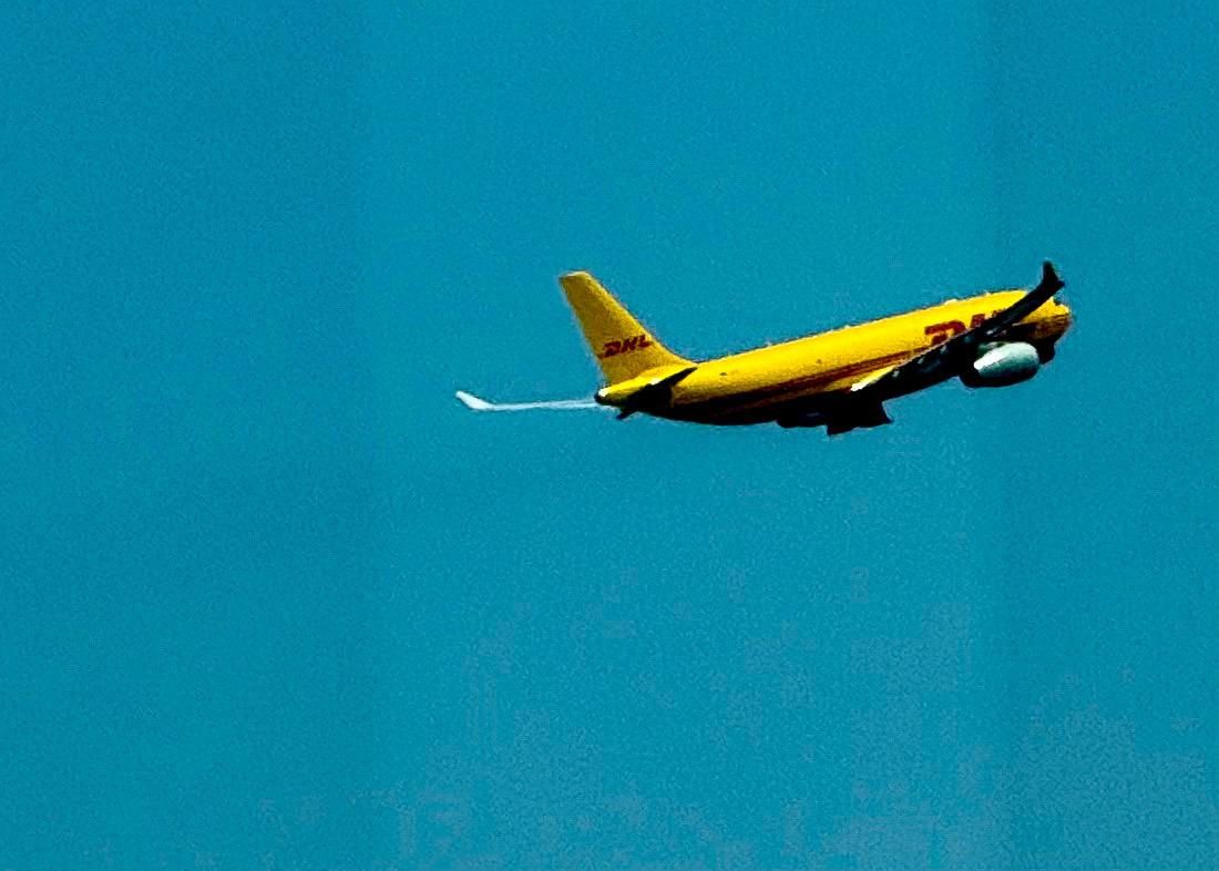 一架黄色的 DHL 快递飞机在蓝天中飞行，喷出白色尾焰。