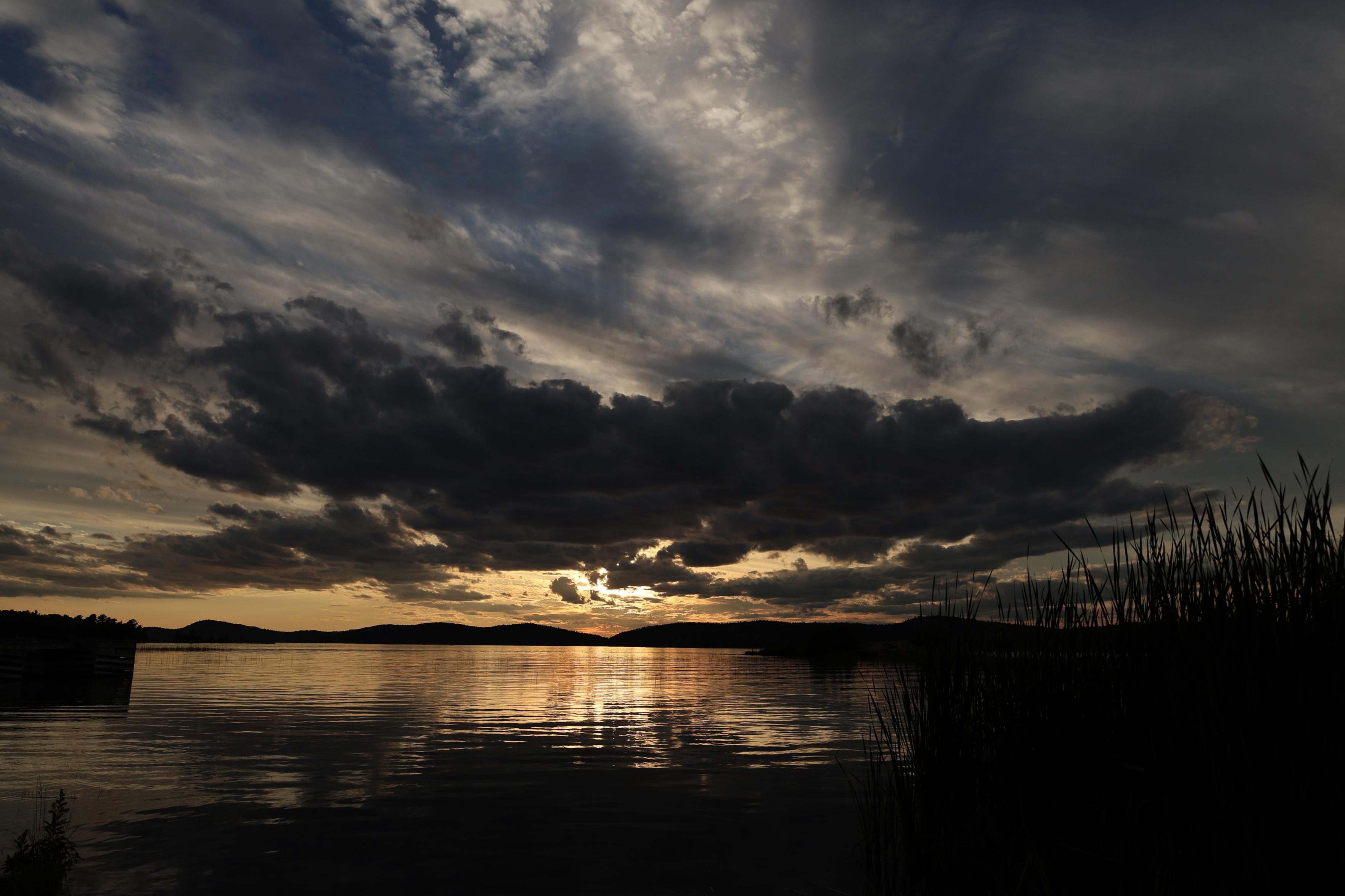 This is a landscape format photo of a summer sunset over a larger lake. The silhouette of a lower level mountain range can be seen in the distance. The sun is partially shrouded in clouds with a very large dark cloud extending across most of the width of the photo. The orange glow from the sun is reflected on the water, creating a shimmering effect. A portion of a raised dock is in the left foreground with taller grasses on the right.