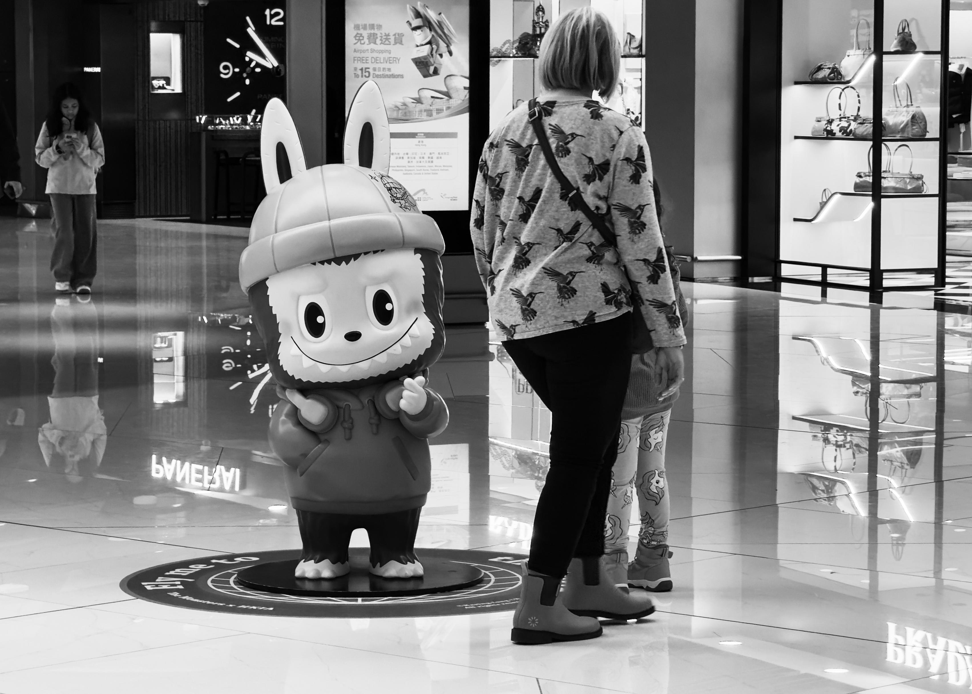 A woman and a child in a shopping mall, standing near a cartoon lion statue with a helmet, surrounded by display cases and signs.