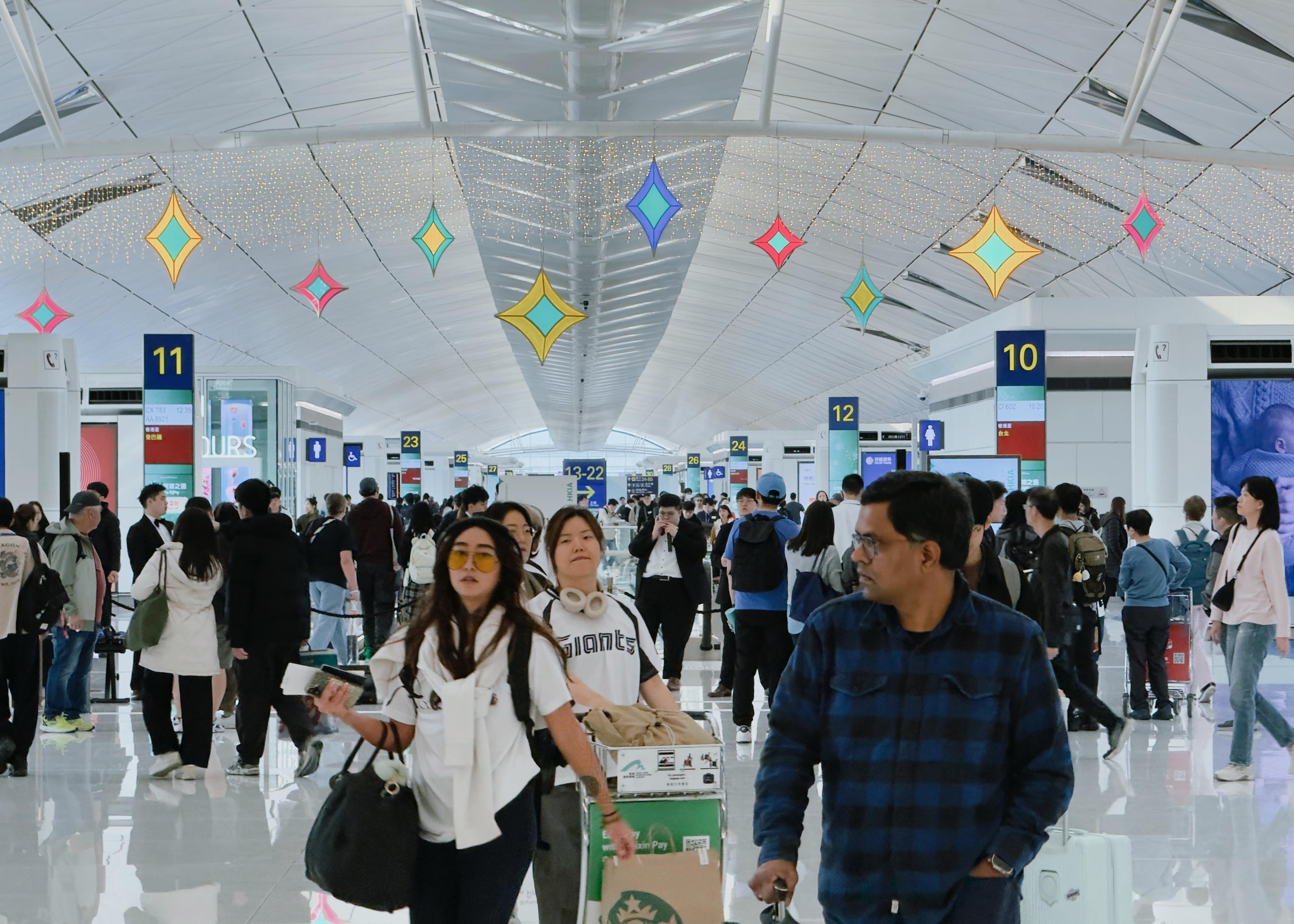 Crowded airport terminal with travelers walking and waiting, colorful hanging decorations, and bright informational signs overhead.