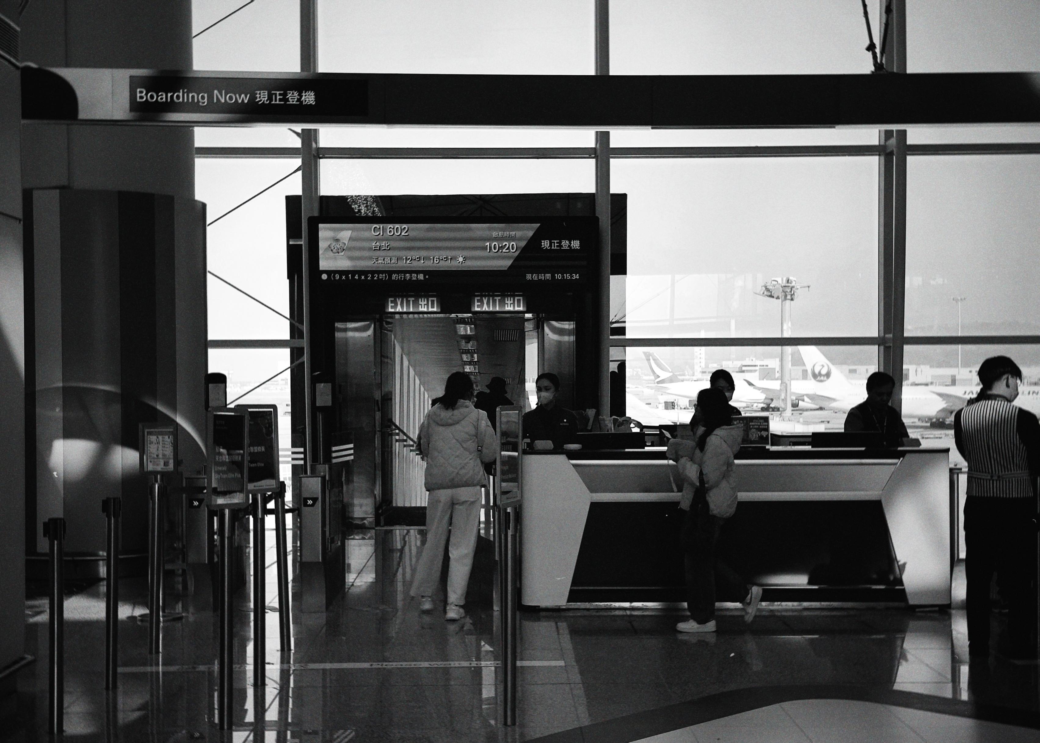Passengers at the airport check-in counter, with planes visible through large windows in the background.