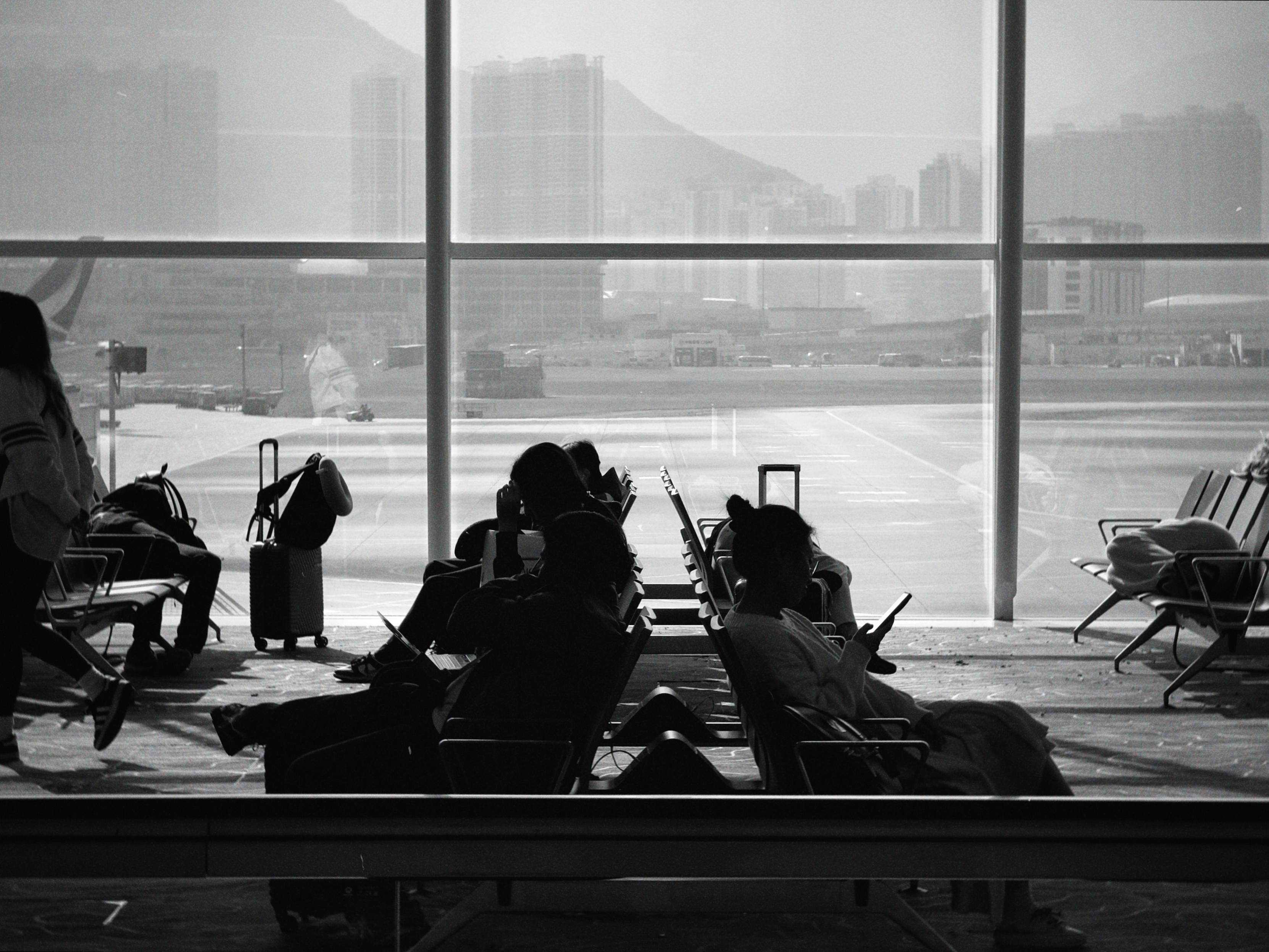 People sitting and waiting at an airport terminal, some using their phones, with large windows showing the runway and cityscape outside.