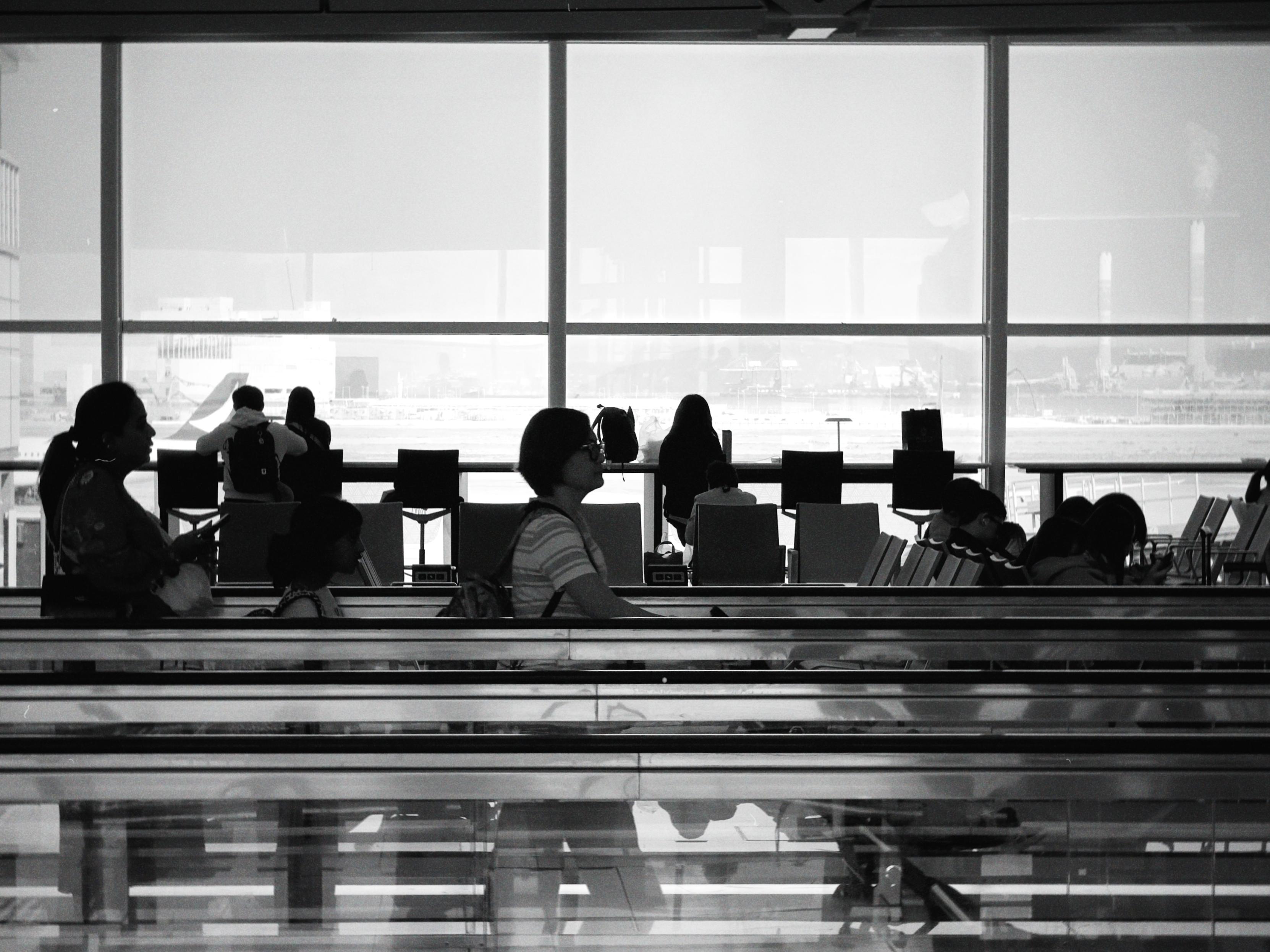 People sitting and standing at an airport terminal with large windows overlooking the runway in the background.