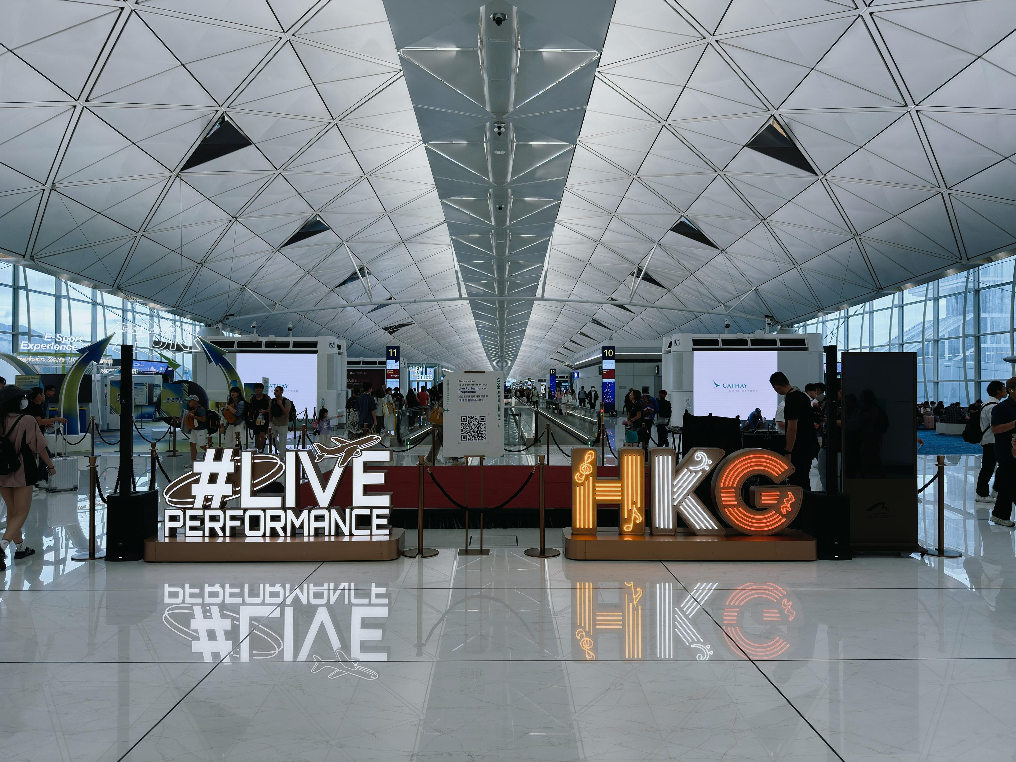 Airport terminal with people walking, large illuminated signs reading "#LIVE PERFORMANCE" and "HKG", and a modern, curved glass ceiling.