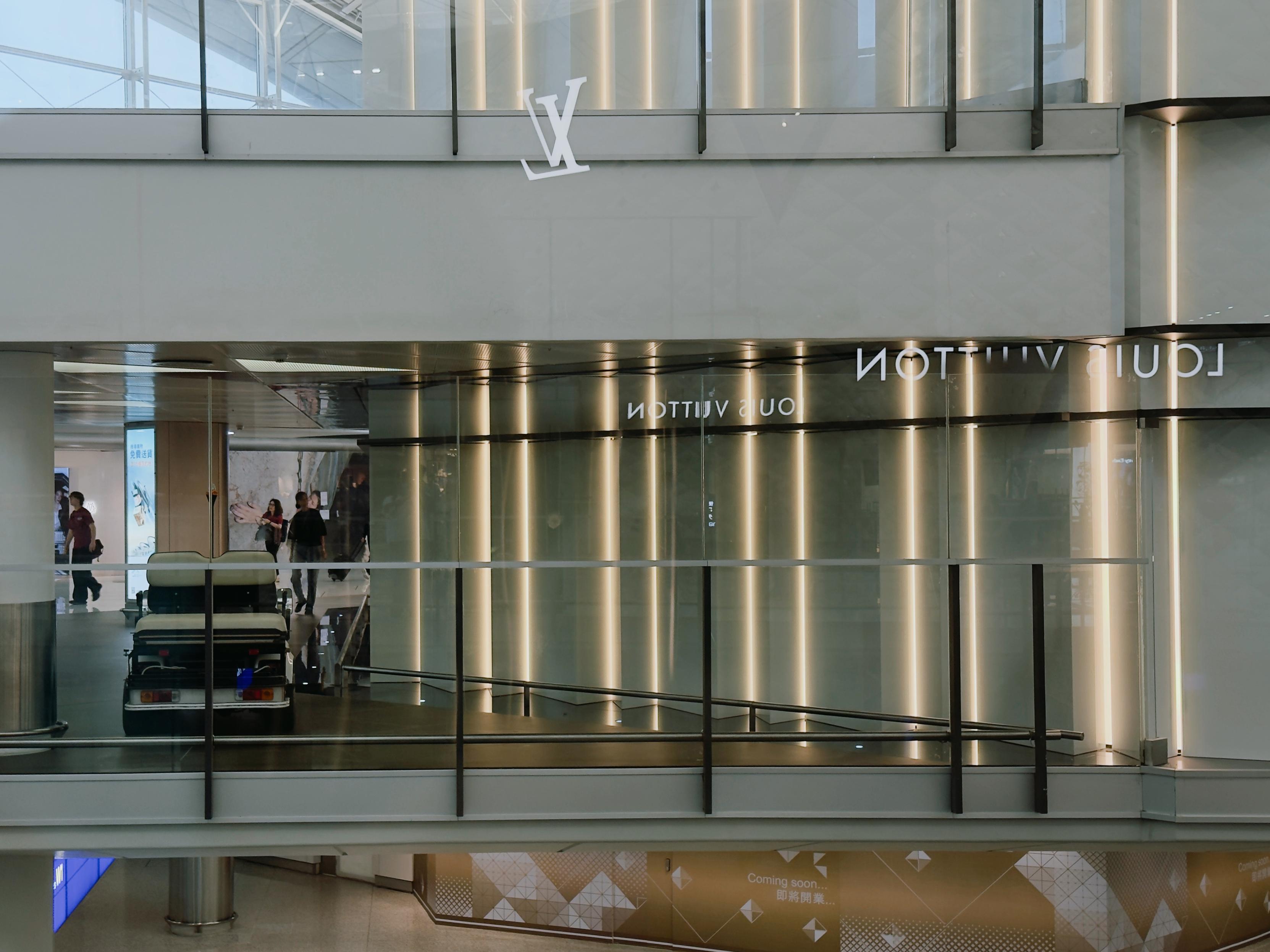 Elevator inside a modern building, reflecting signage such as "Louis Vuitton" on glass walls, with a few people walking in the background.