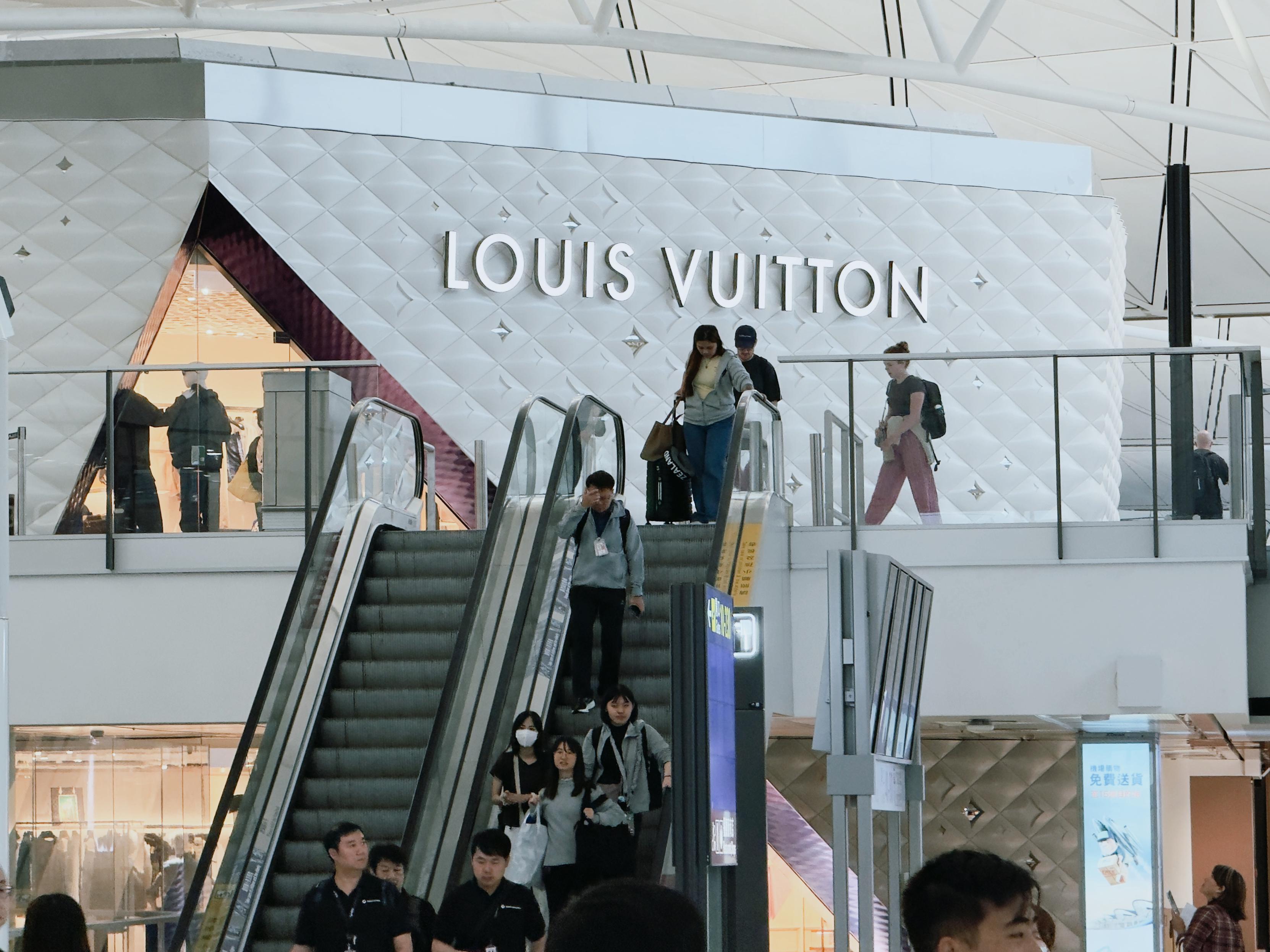 People walking up and down escalators inside a Louis Vuitton store at an airport.