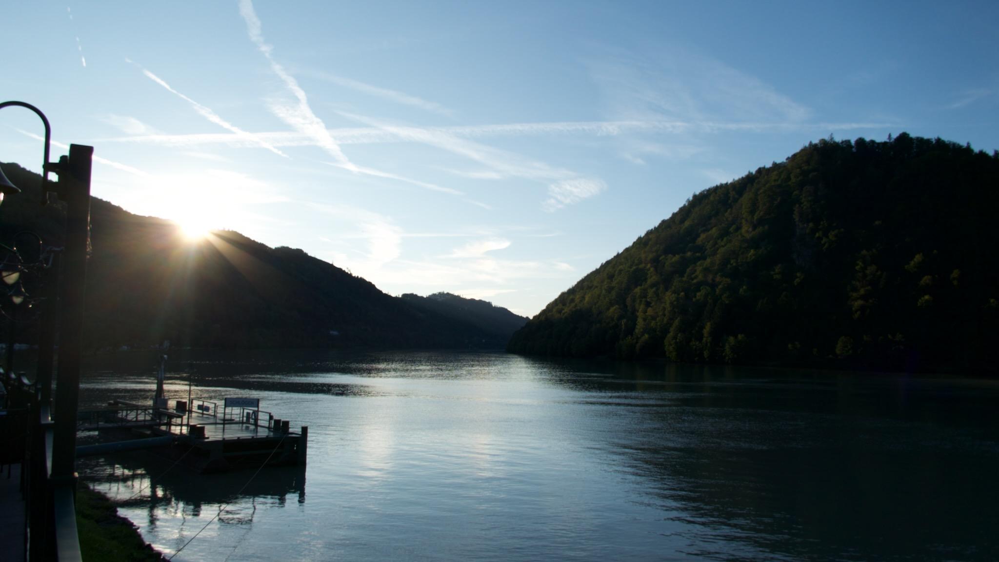Picture of the river Danube in Upper Austria. The river is in the middle of the photo, clearly the subject. on the right side of the water is a gentle hill in darkness as it is evening. on the left side the hill is not that steep and high as on the opposite. On the left side the sun is glimpsing with sunbeams and flares. 
The upper half and background is a blue sky with some clouds. In the forefront there is a ship landing construction in dark shadows.